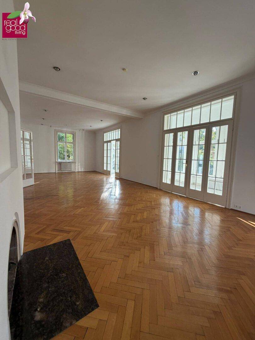 Großzügiges Wohnzimmer mit Fischgrätparkett, hohen Fenstertüren und einem eleganten Kamin.