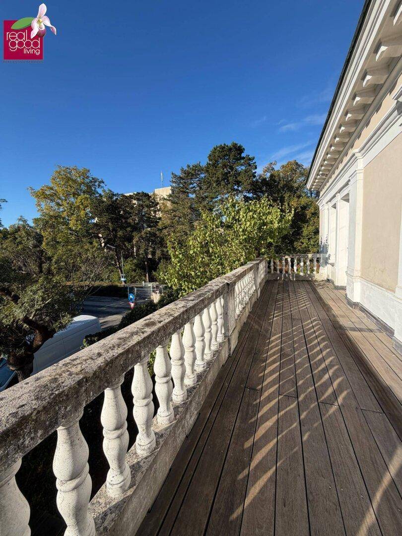 Langer Balkon mit Holzplanken und klassischer Balustrade, umgeben von grünen Bäumen.