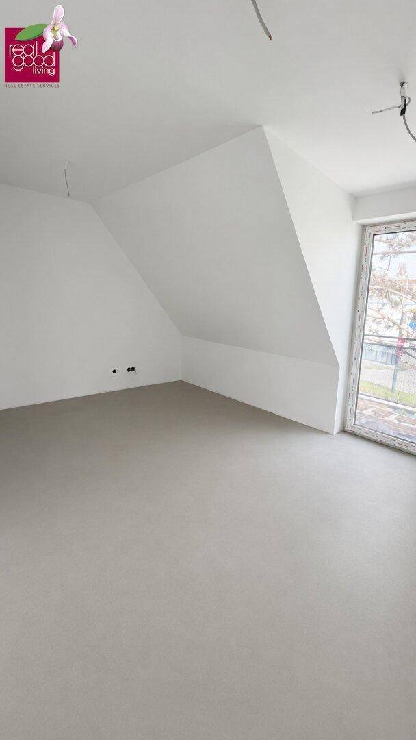 Heller, unmöblierter Raum mit schrägen Wänden, großem Fenster und hellem Bodenbelag.