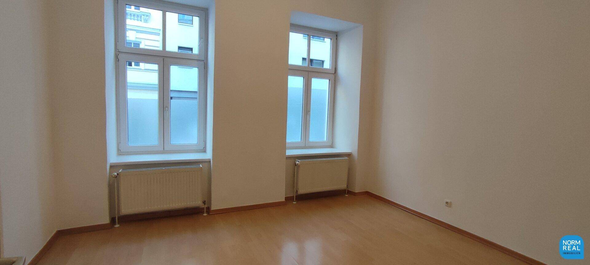 Geräumiges Zimmer mit viel Tageslicht durch zwei große Fenster und hellem Holzfußboden.