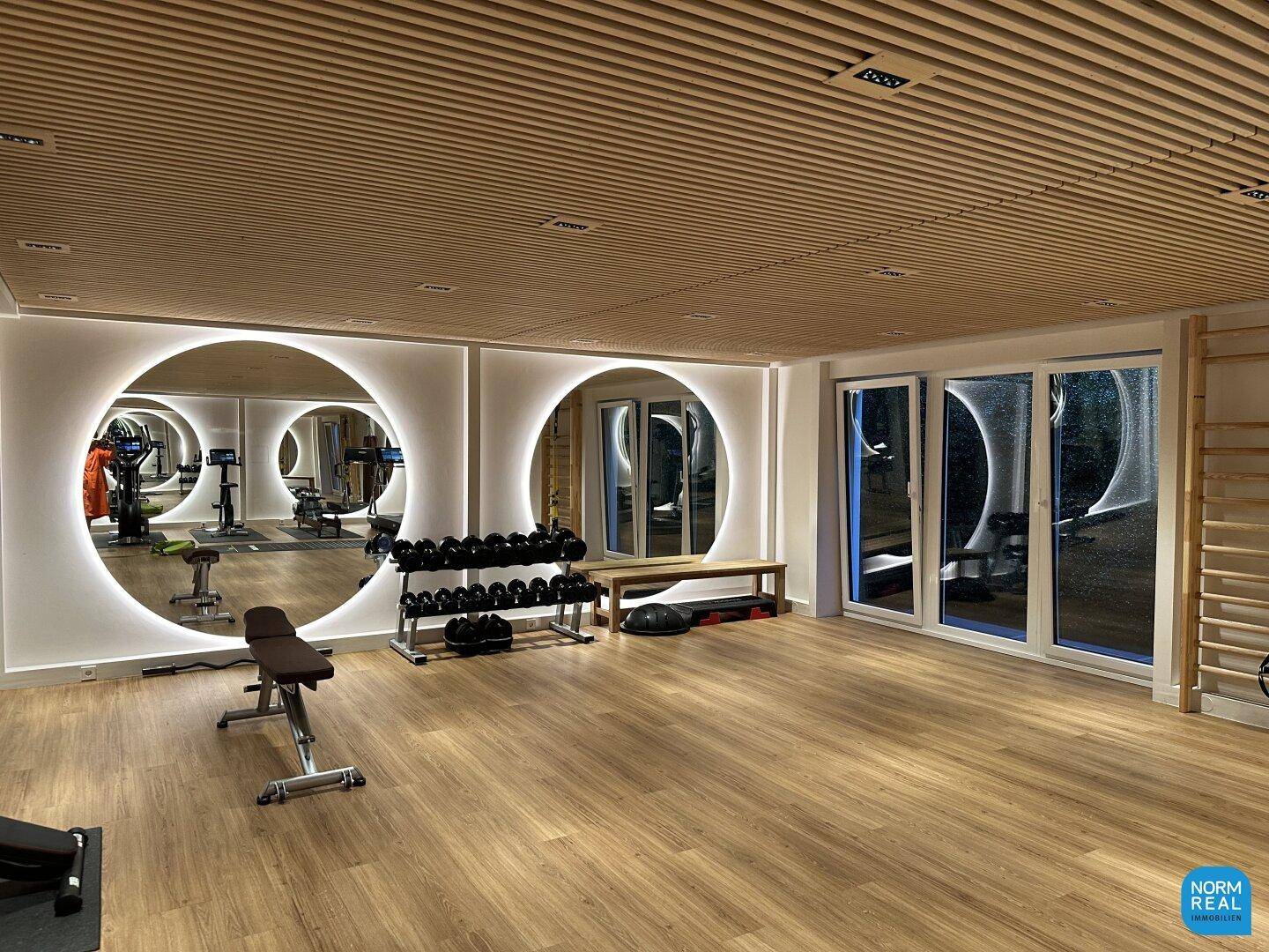 Gut ausgestatteter Fitnessraum mit Holzlamellendecke, großen Spiegeln und Trainingsgeräten.