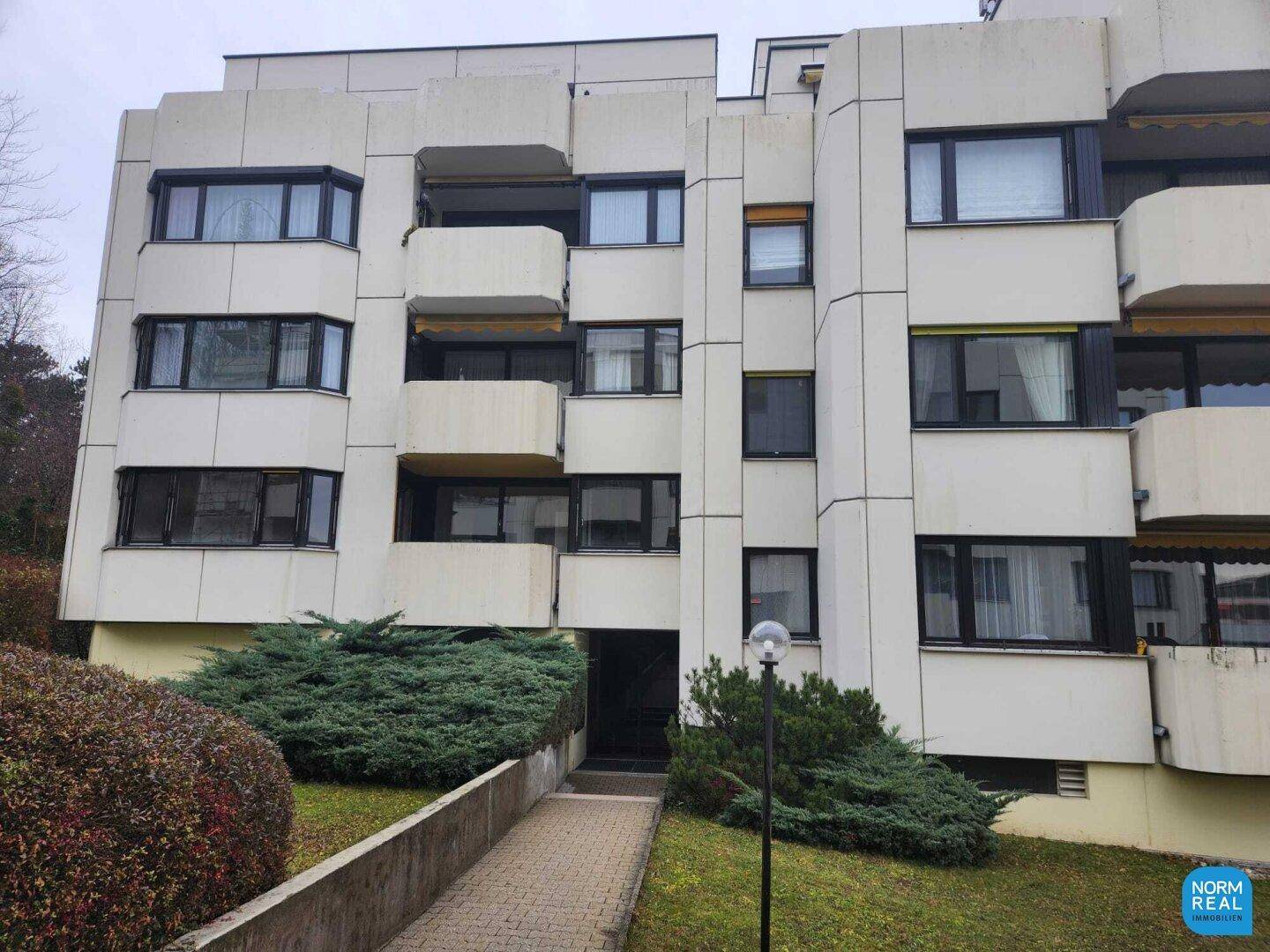 Mehrfamilienhaus mit mehreren Balkonen und großen Fenstern in einer grünen Umgebung.