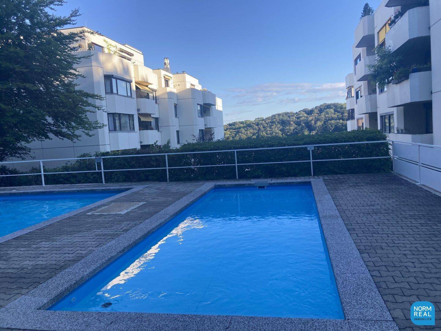 Außenansicht der Wohnanlage mit zwei Pools und Blick auf die umliegende grüne Hügellandschaft.