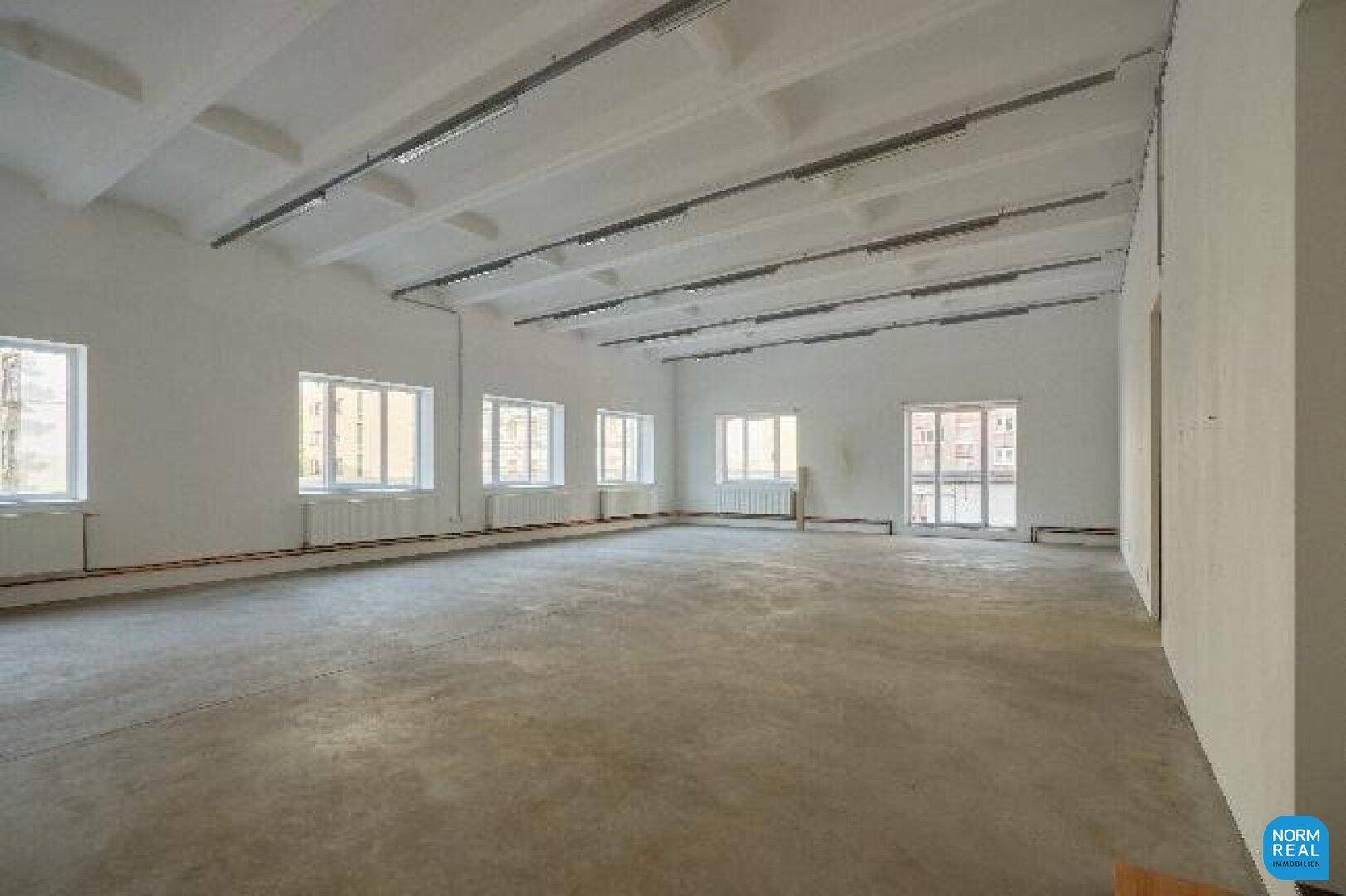 Weitläufiger, heller Raum mit vielen Fenstern, Betonboden und industrieller Deckenbeleuchtung.
