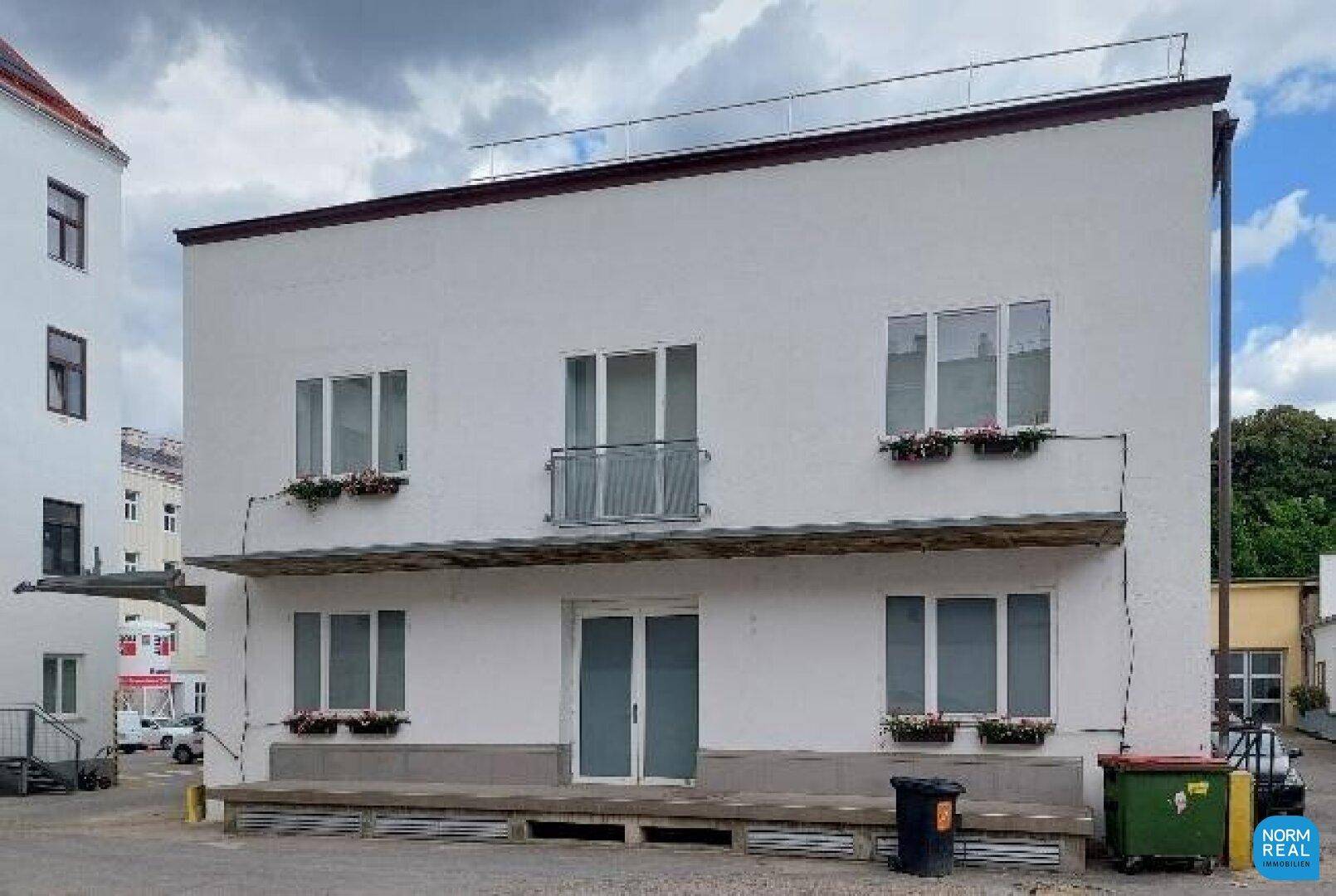 Weißes Gebäude mit mehreren Fenstern, Blumenkästen und einem kleinen Balkon unter bewölktem Himmel.