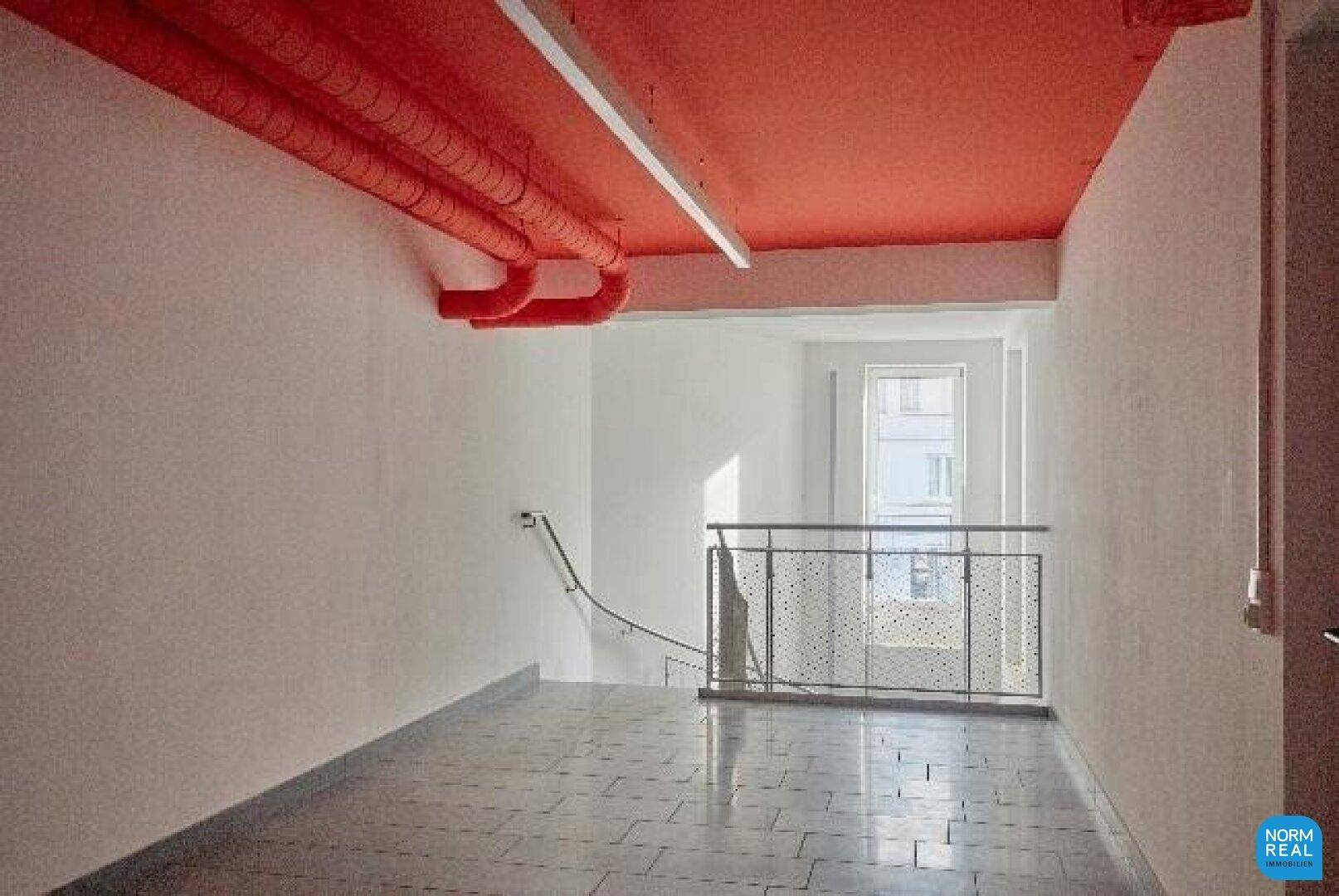 Flur mit roter Decke, sichtbaren Rohren und Treppenabgang mit Metallgeländer.
