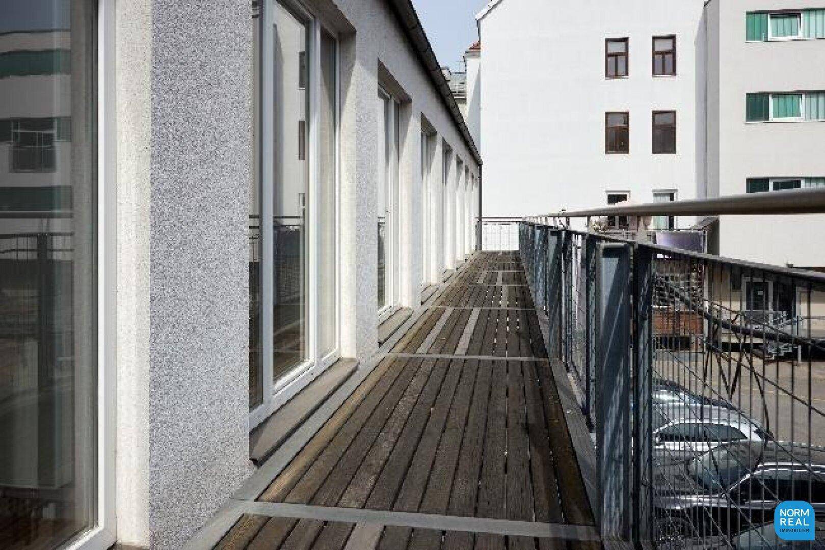 Langer Balkon mit Holzdielen und Metallgeländer, bietet Zugang zu mehreren Räumen und Blick auf die Umgebung.