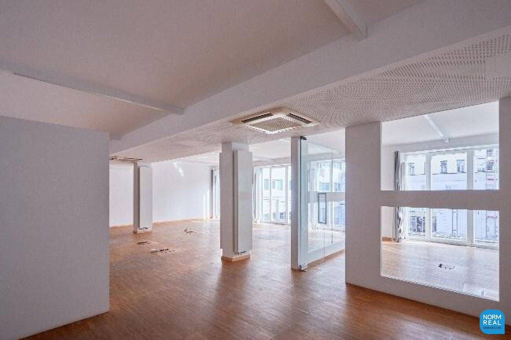 Großzügiger Büroraum mit Holzboden, vielen Fenstern und integrierter Klimaanlage, ideal für flexible Nutzung.