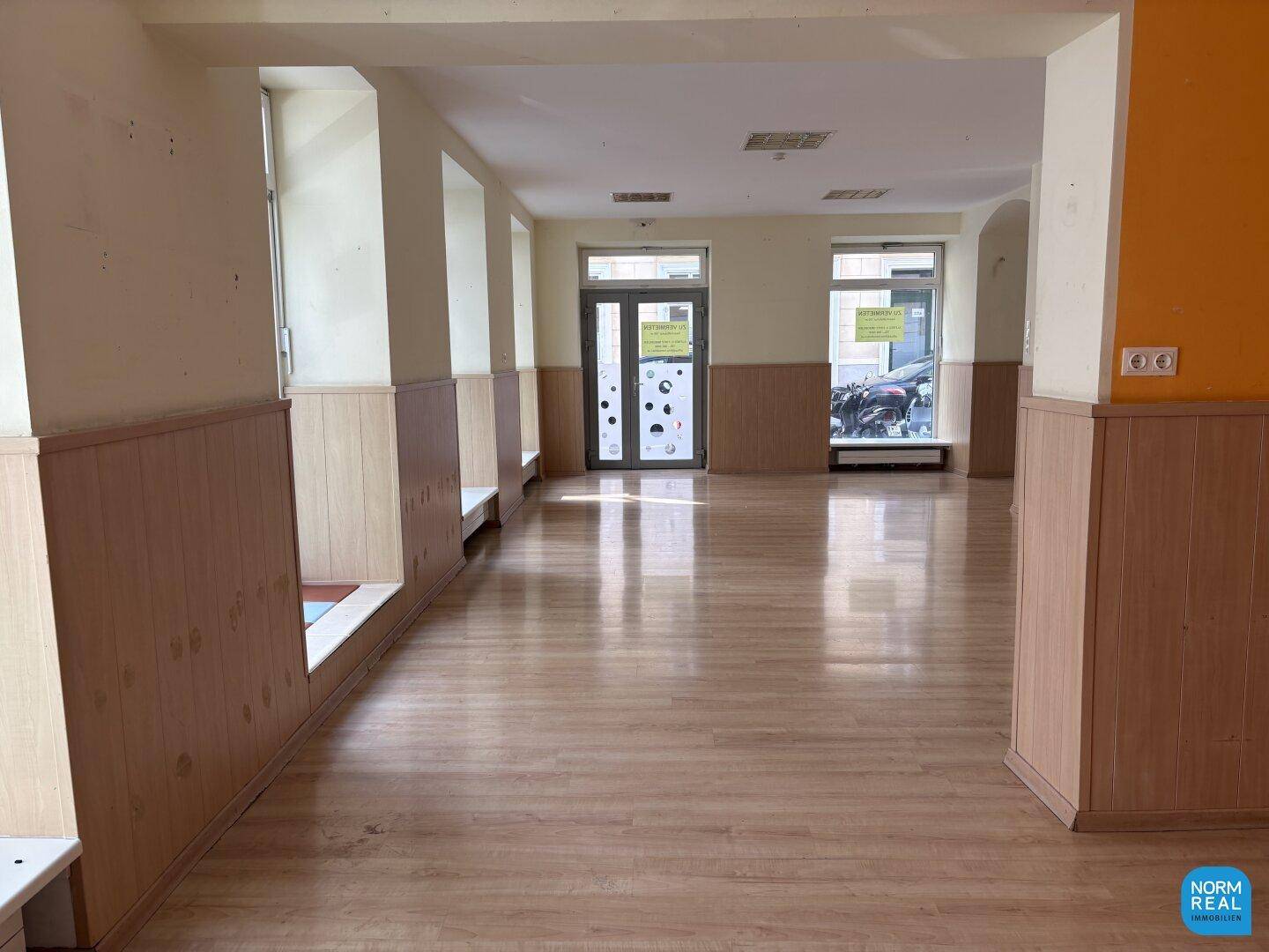 Heller Gewerberaum mit Laminatboden und großen Schaufenstern, die viel Tageslicht hereinlassen.