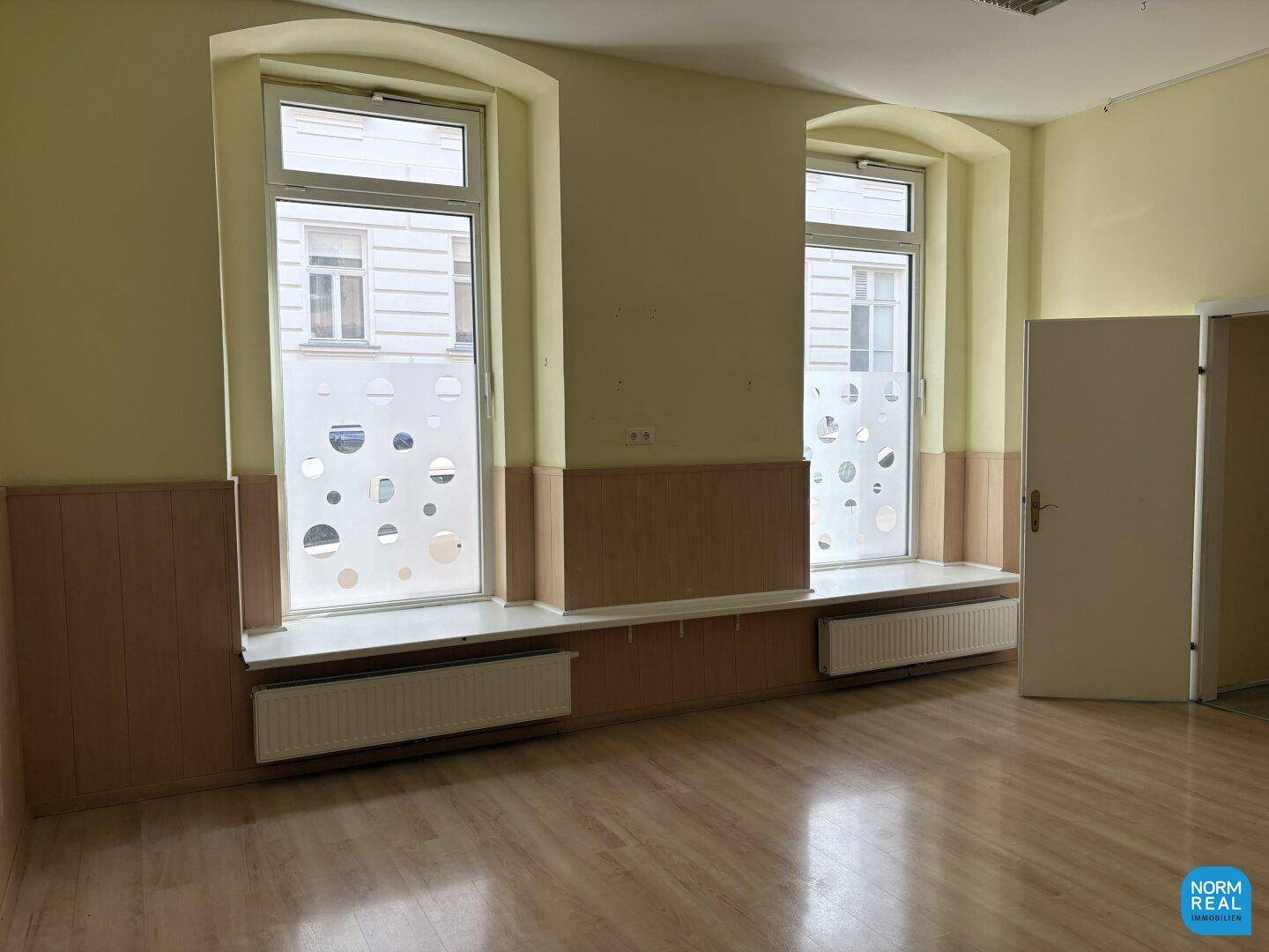 Gewerberaum mit zwei großen Fenstern, Heizkörpern und Holzpaneelen an den Wänden.