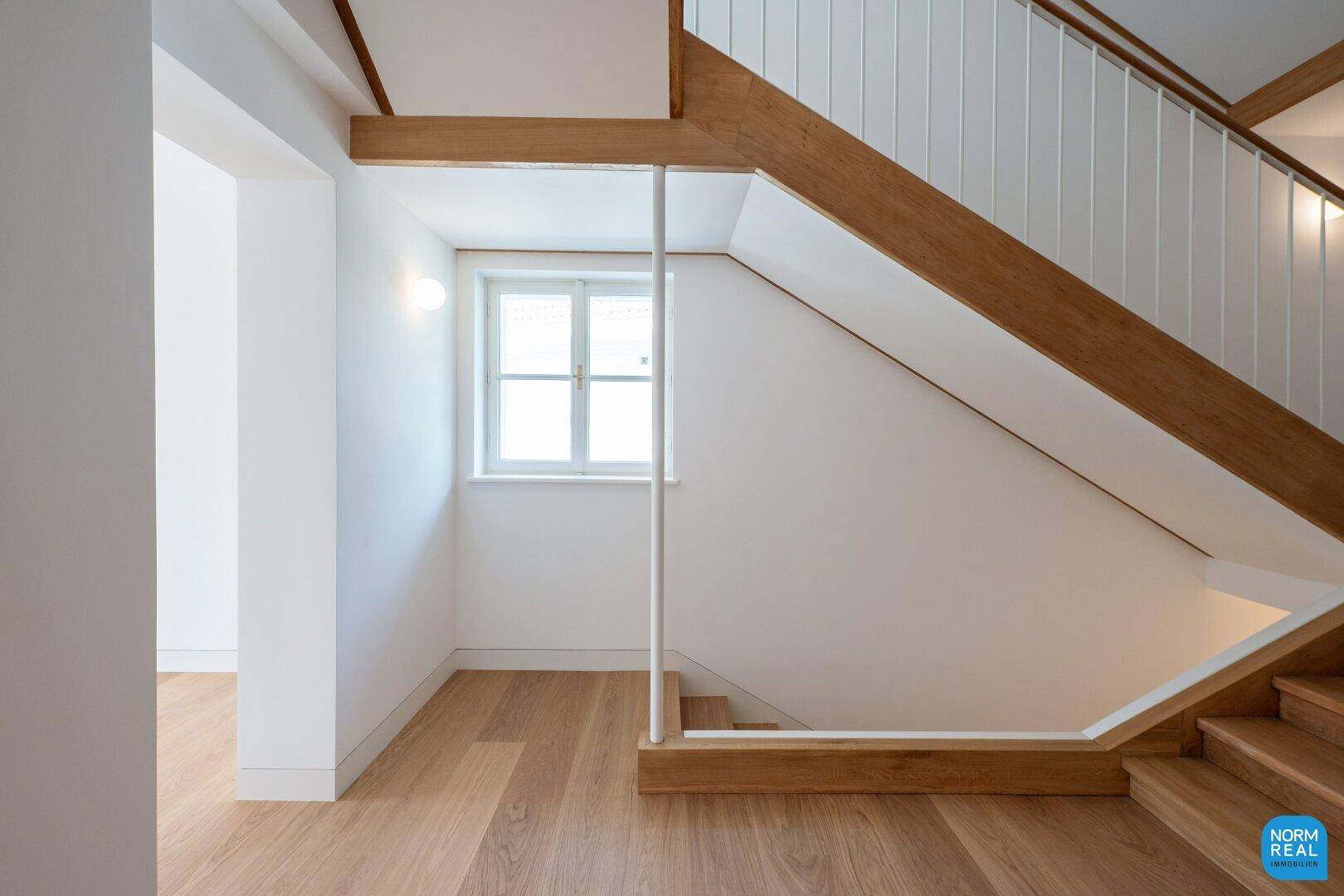 Helles Treppenhaus mit Holzstufen, weißen Geländern und einem Fenster für natürliches Licht.