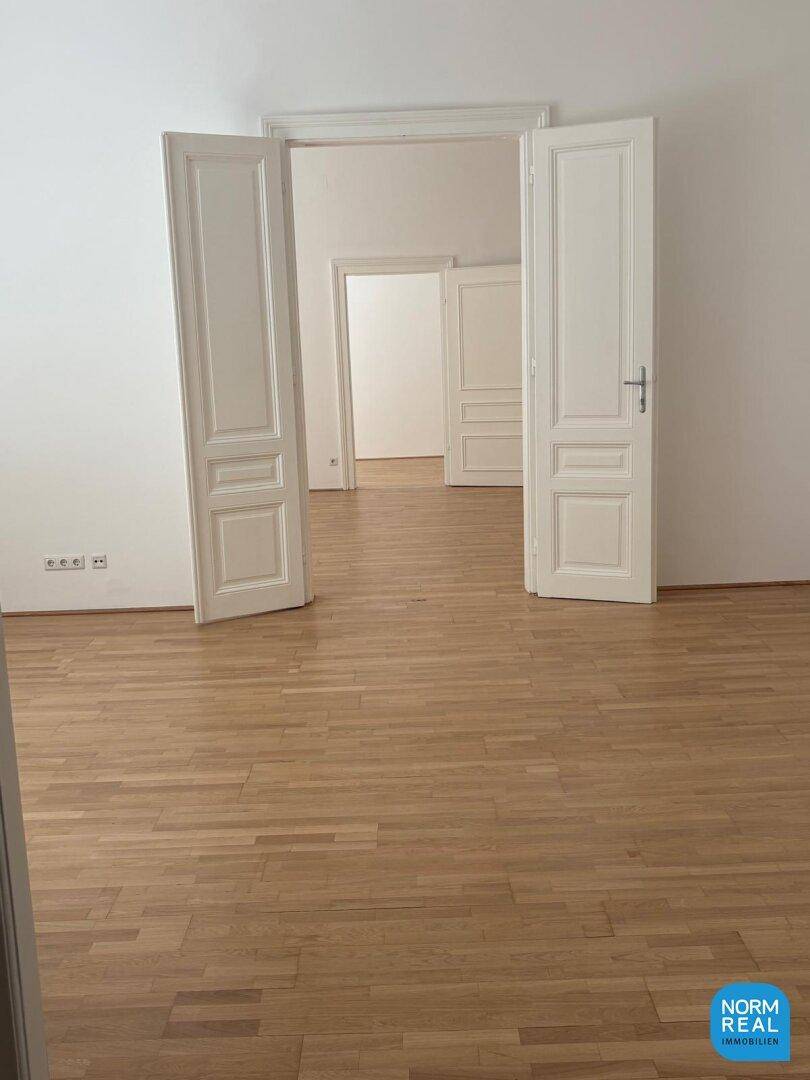 Heller Raum mit Fischgrätparkett und hohen Türen, der in weitere Zimmer führt und viel Potenzial bietet.