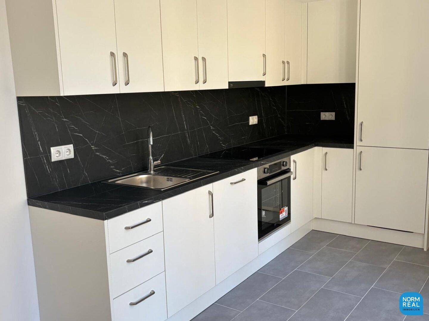 Moderne Einbauküche mit weißen Schränken, schwarzen Arbeitsplatten und integrierten Geräten auf grauem Fliesenboden.
