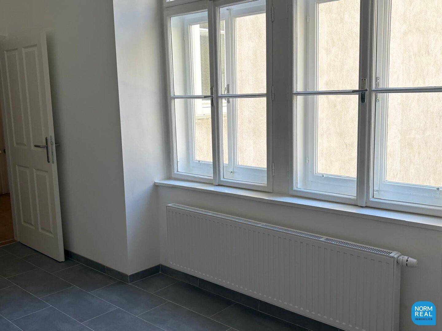 Raum mit grauen Fliesen und großem Fenster, bereit für die Kücheninstallation oder als Hauswirtschaftsraum nutzbar.
