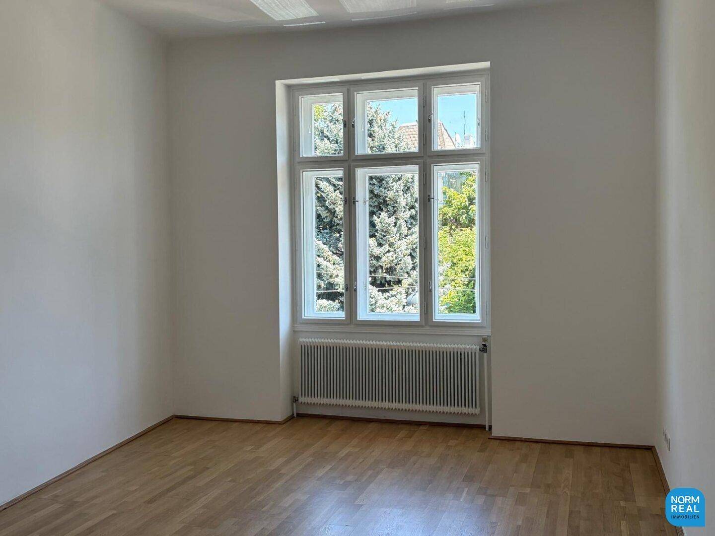 Heller Raum mit Parkettboden und einem großen Fenster, das einen schönen Blick ins Grüne bietet und den Raum erhellt.