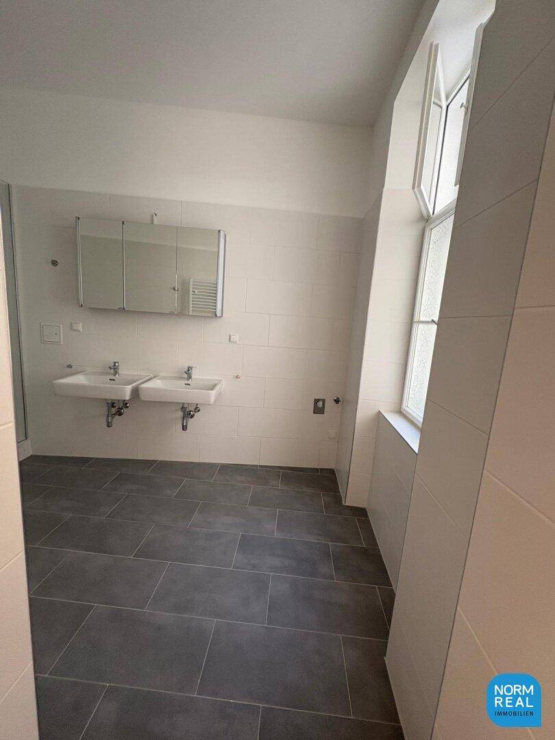 Helles, modernes Badezimmer mit Doppelwaschbecken, Spiegelschrank und großem Fenster, das viel Licht spendet.