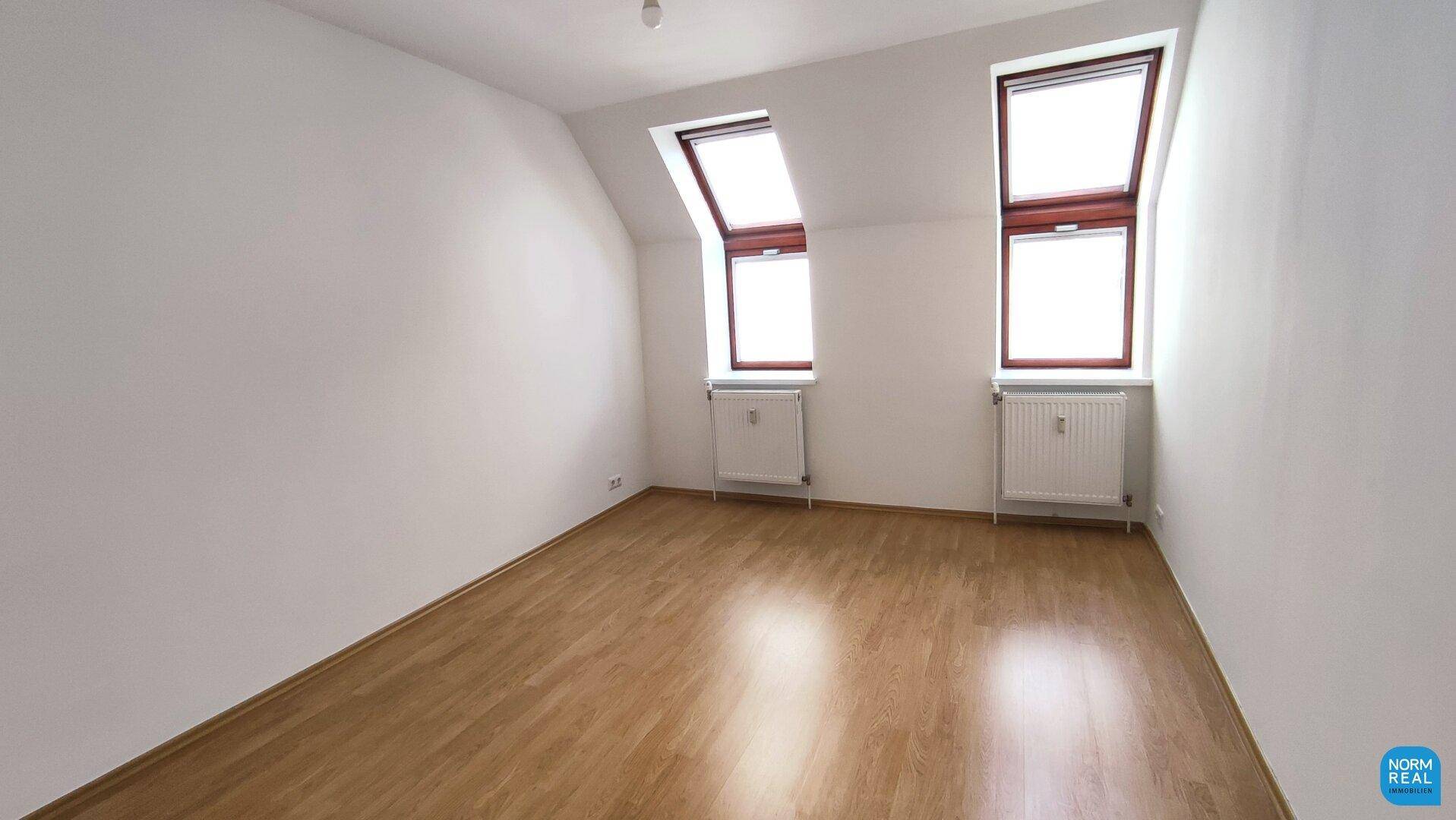 Helles Schlafzimmer mit Holzboden, zwei Dachfenstern und Heizkörpern unter der Dachschräge.