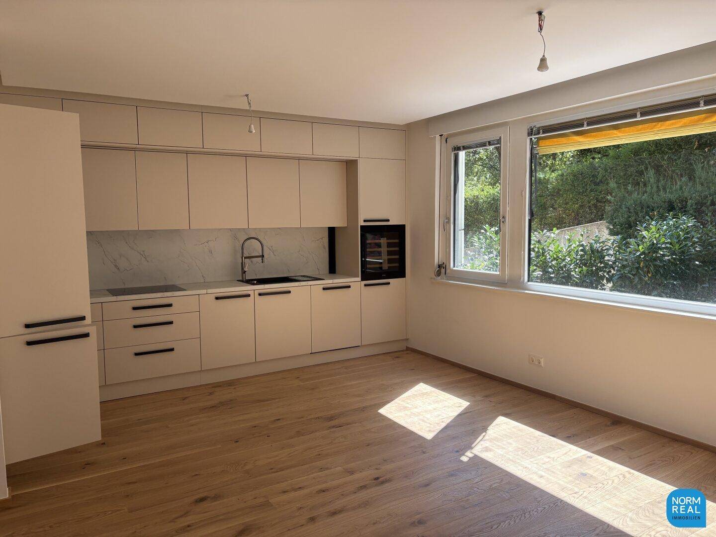 Moderne Einbauküche mit hellen Fronten, Holzboden und Blick ins Grüne durch große Fenster.