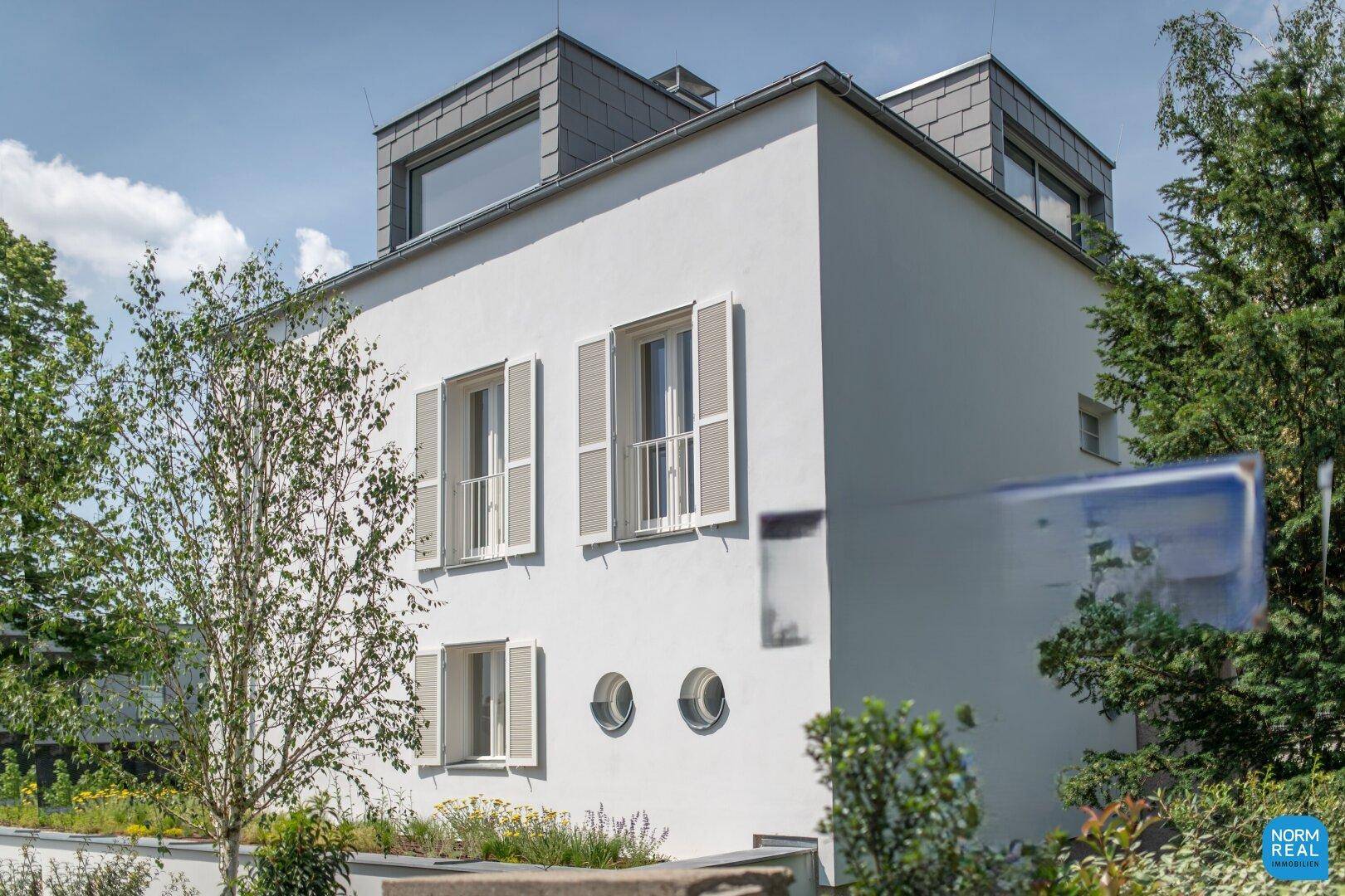 Modernes Wohnhaus mit weißer Fassade, Dachgauben und gepflegter Bepflanzung im Vordergrund.