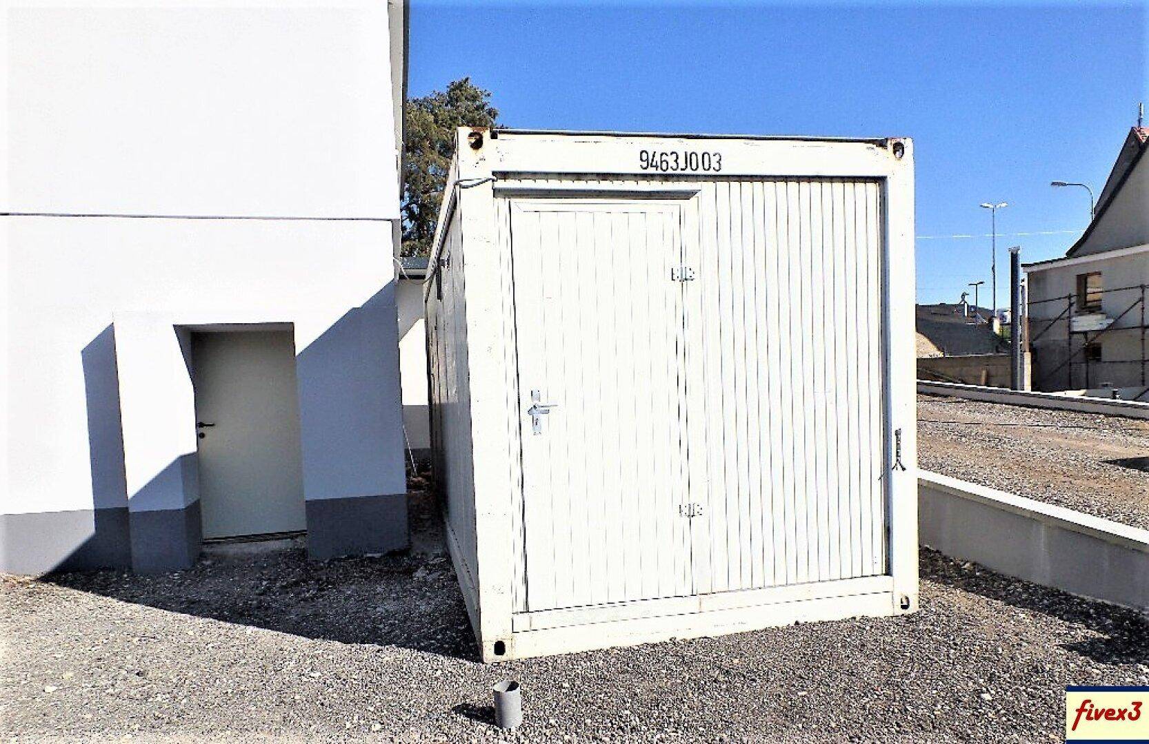 Ein weißer Lagercontainer steht neben einem Gebäude auf einem Schotterplatz unter klarem blauem Himmel.