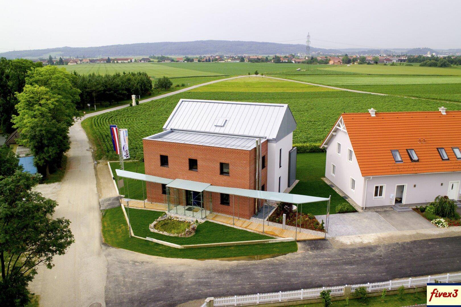 Luftaufnahme eines modernen Backsteinhauses mit Carport und gepflegter Grünfläche, umgeben von Feldern.