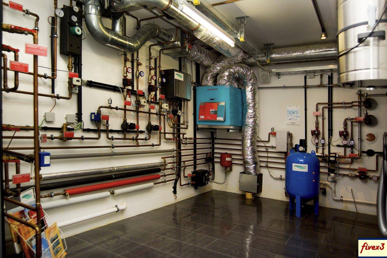 Technikraum mit komplexen Rohrleitungen, Heizungsanlage und einem blauen Druckbehälter.