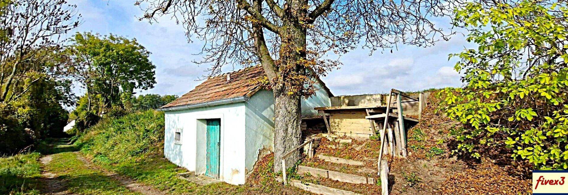 Panoramablick auf ein kleines, altes Gebäude in ländlicher Umgebung mit einem Weg und Bäumen unter blauem Himmel.