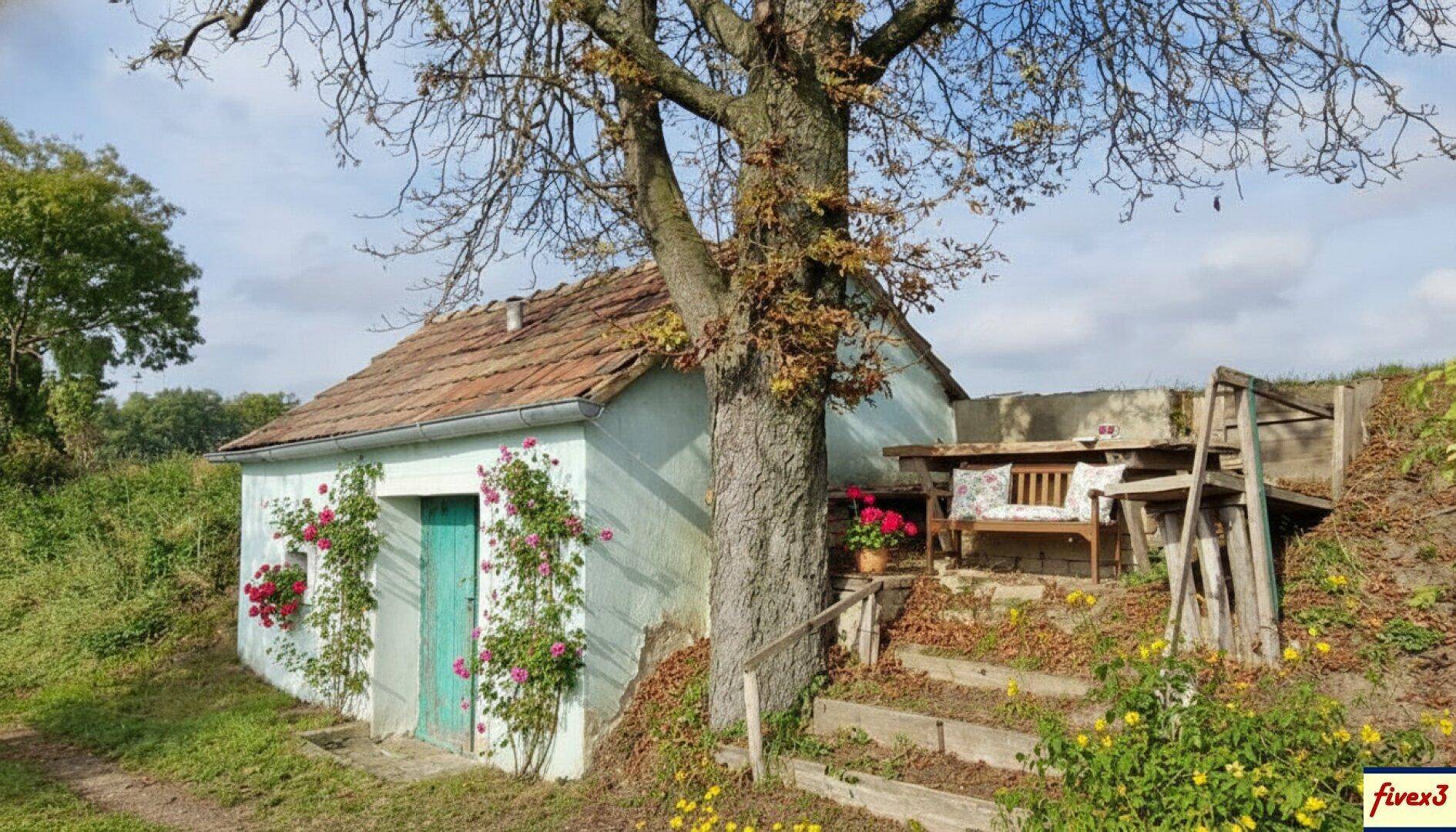 Kleines, altes Gebäude mit Ziegeldach und einem Baum davor, umgeben von Grün und einer rustikalen Sitzecke.