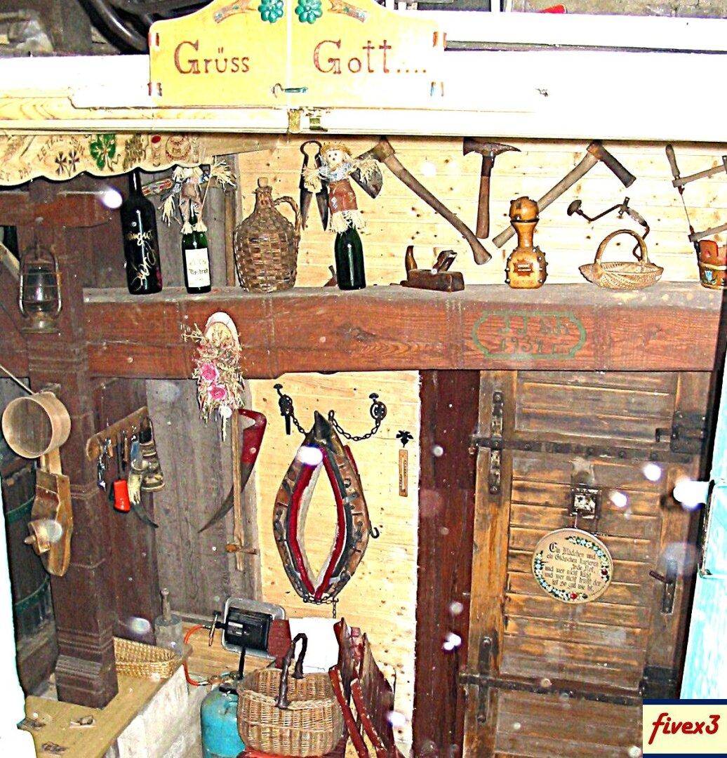 Rustikale Holzwand mit traditionellen Werkzeugen und Dekorationen, darunter ein Schild mit der Aufschrift "Grüss Gott".