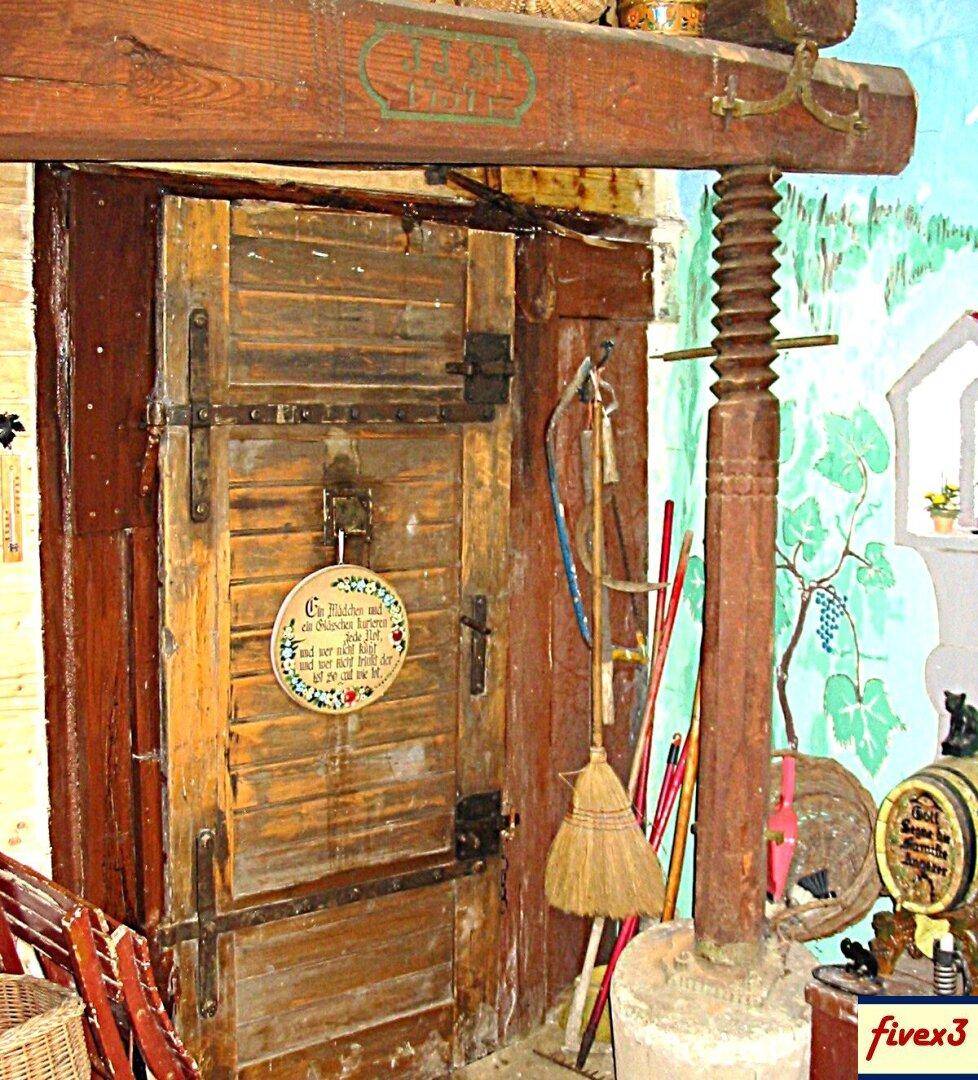 Rustikale Holztür mit Metallbeschlägen neben einer alten Weinpresse und einer Wandmalerei im traditionellen Stil.
