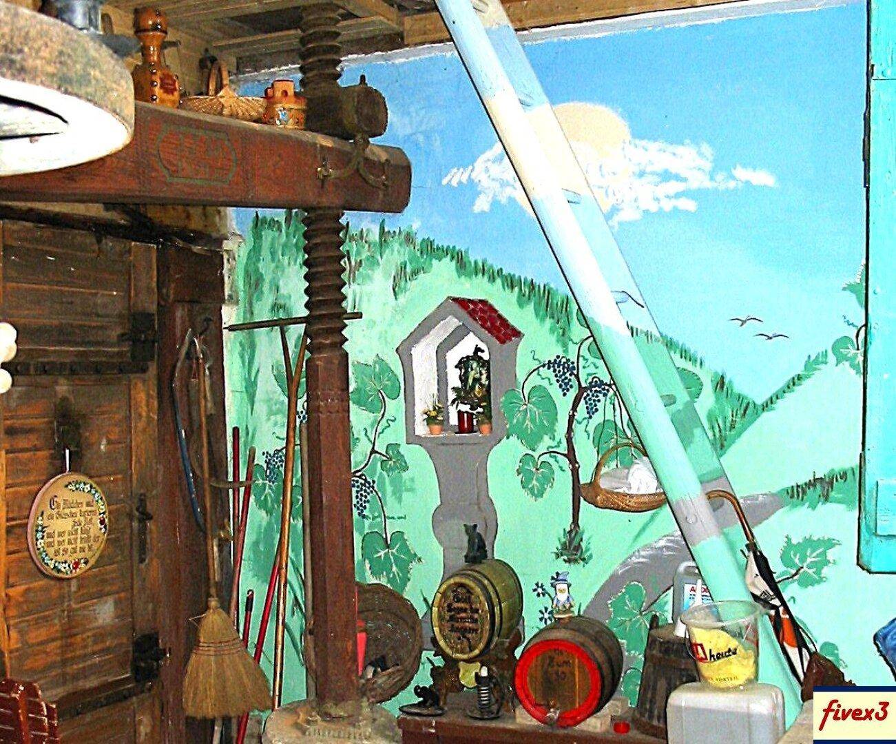 Rustikaler Innenraum mit einer alten Weinpresse, Holzfässern und einer Wandmalerei, die eine Weinlandschaft darstellt.