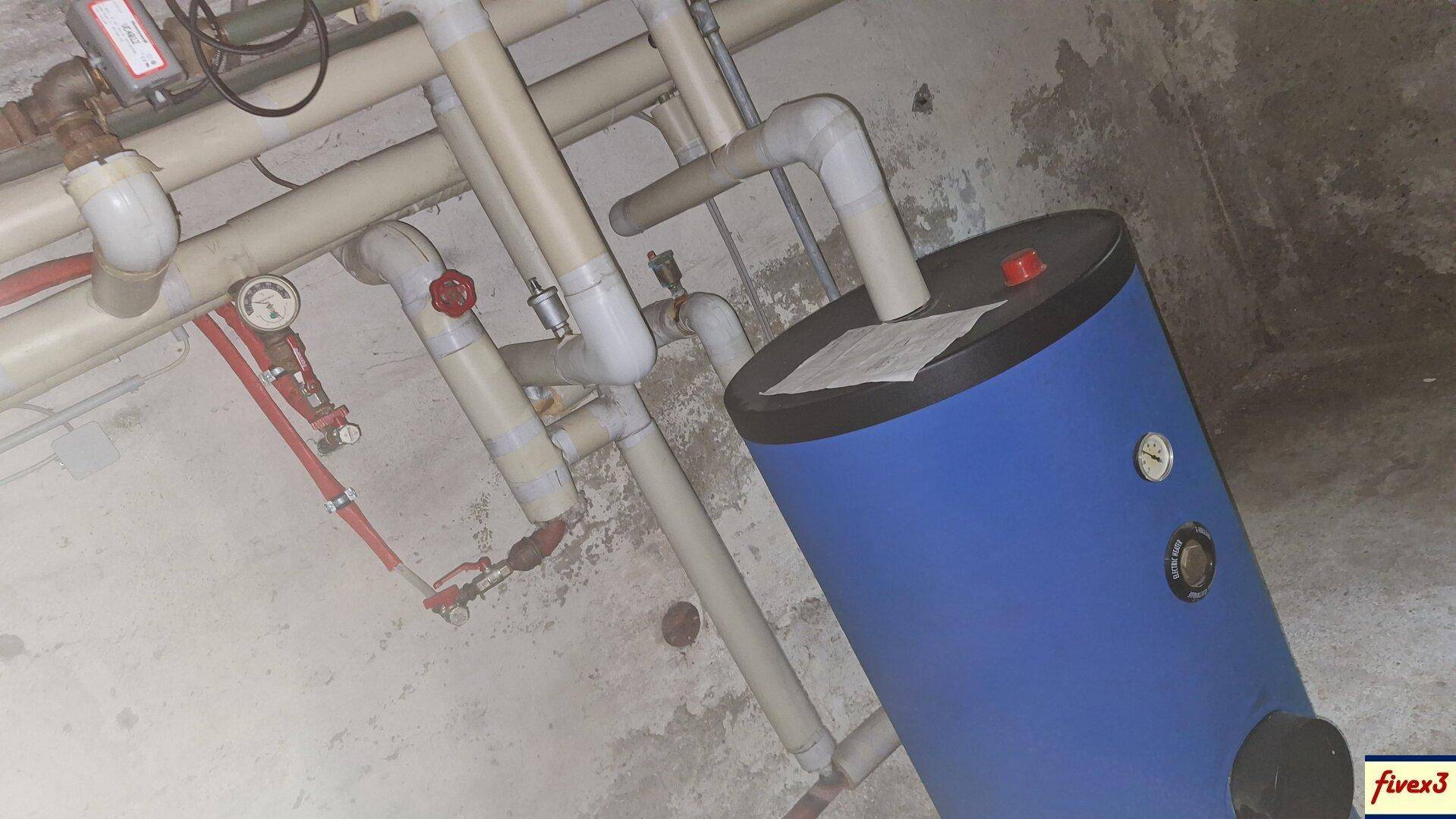 Kellerraum mit sichtbaren Rohren und einem blauen Warmwasserspeicher an der Betonwand.