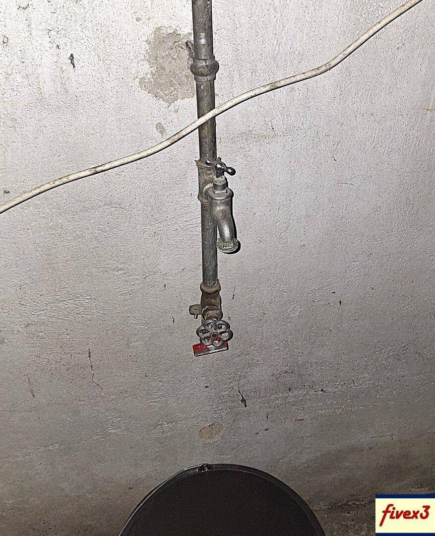 Wasserhahn und Rohrleitungen in einem einfachen Kellerraum mit rauen Wänden.