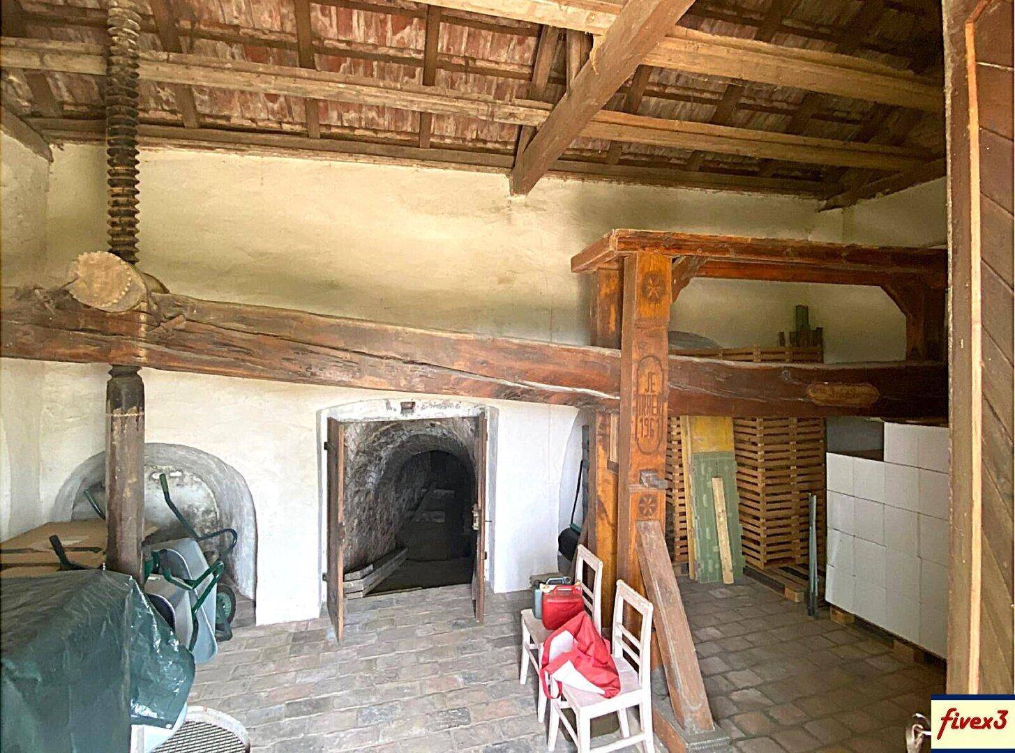 Innenansicht eines rustikalen Kellers mit Holzbalken, Ziegelboden und einem Eingang zu einem weiteren Gewölbe.