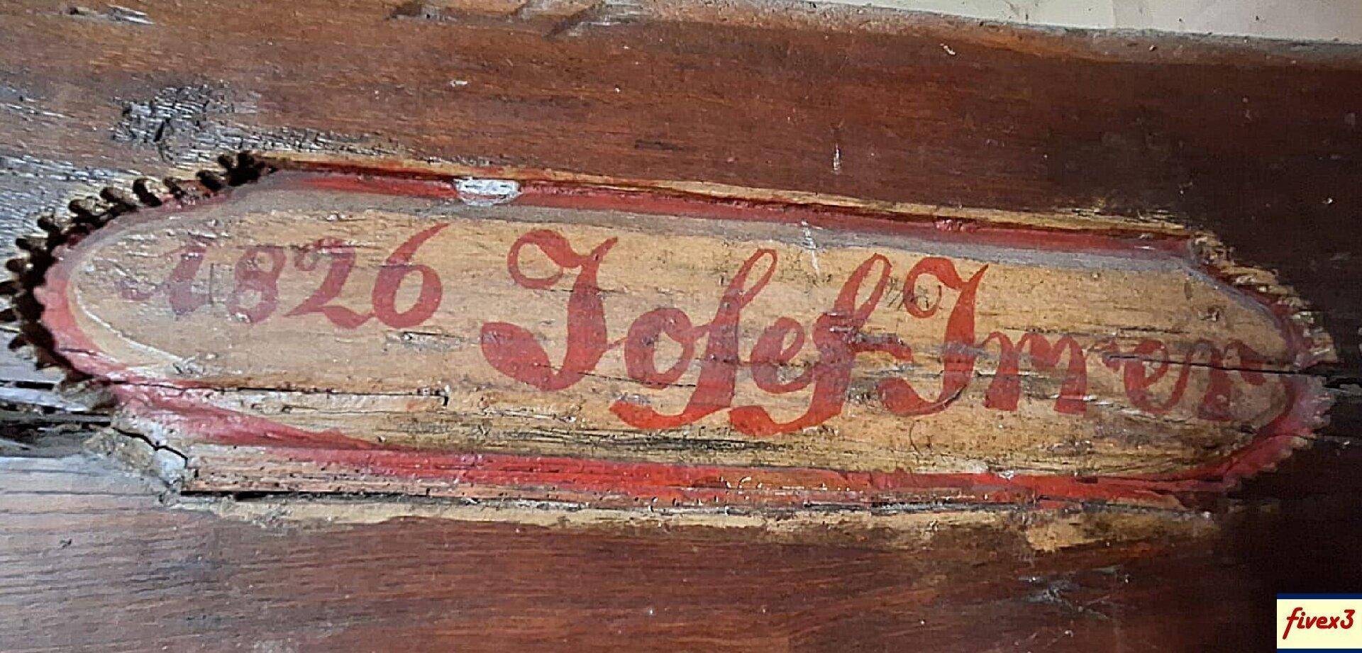 Nahaufnahme eines alten, handbemalten Holzschildes mit der Jahreszahl 1826 und einem Namen.