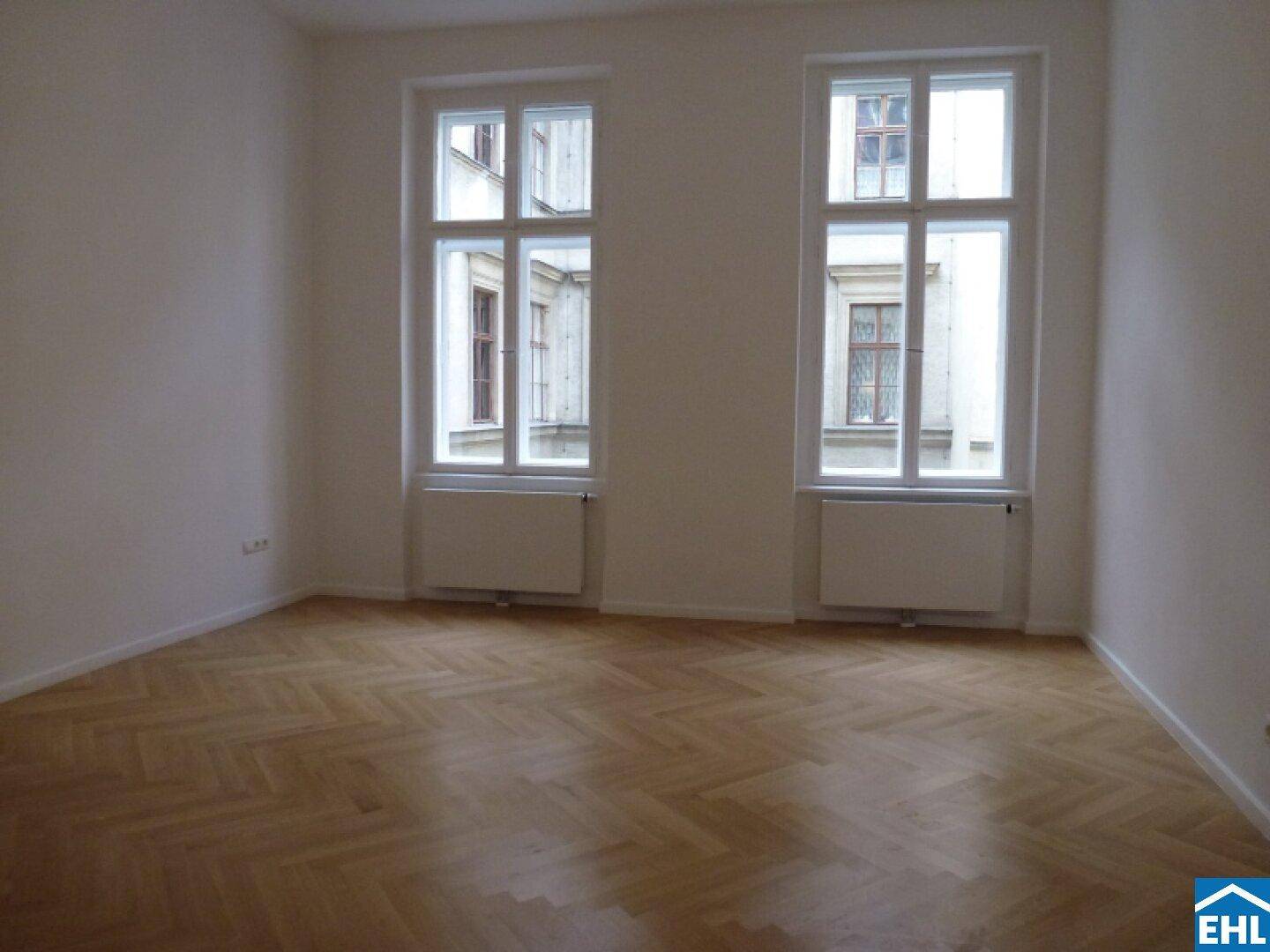 Helles Zimmer mit Fischgrätparkett und zwei hohen Fenstern mit Blick auf den Innenhof.