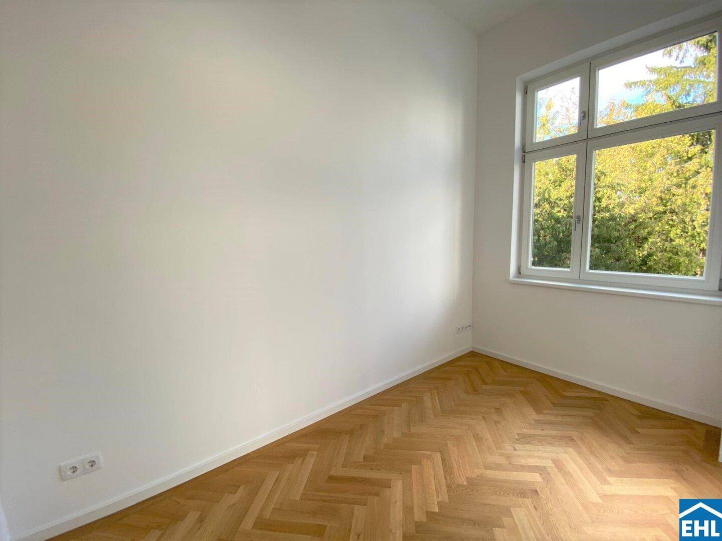 Leeres Zimmer mit Fischgrätparkett und einem großen Fenster, das einen Blick ins Grüne bietet.