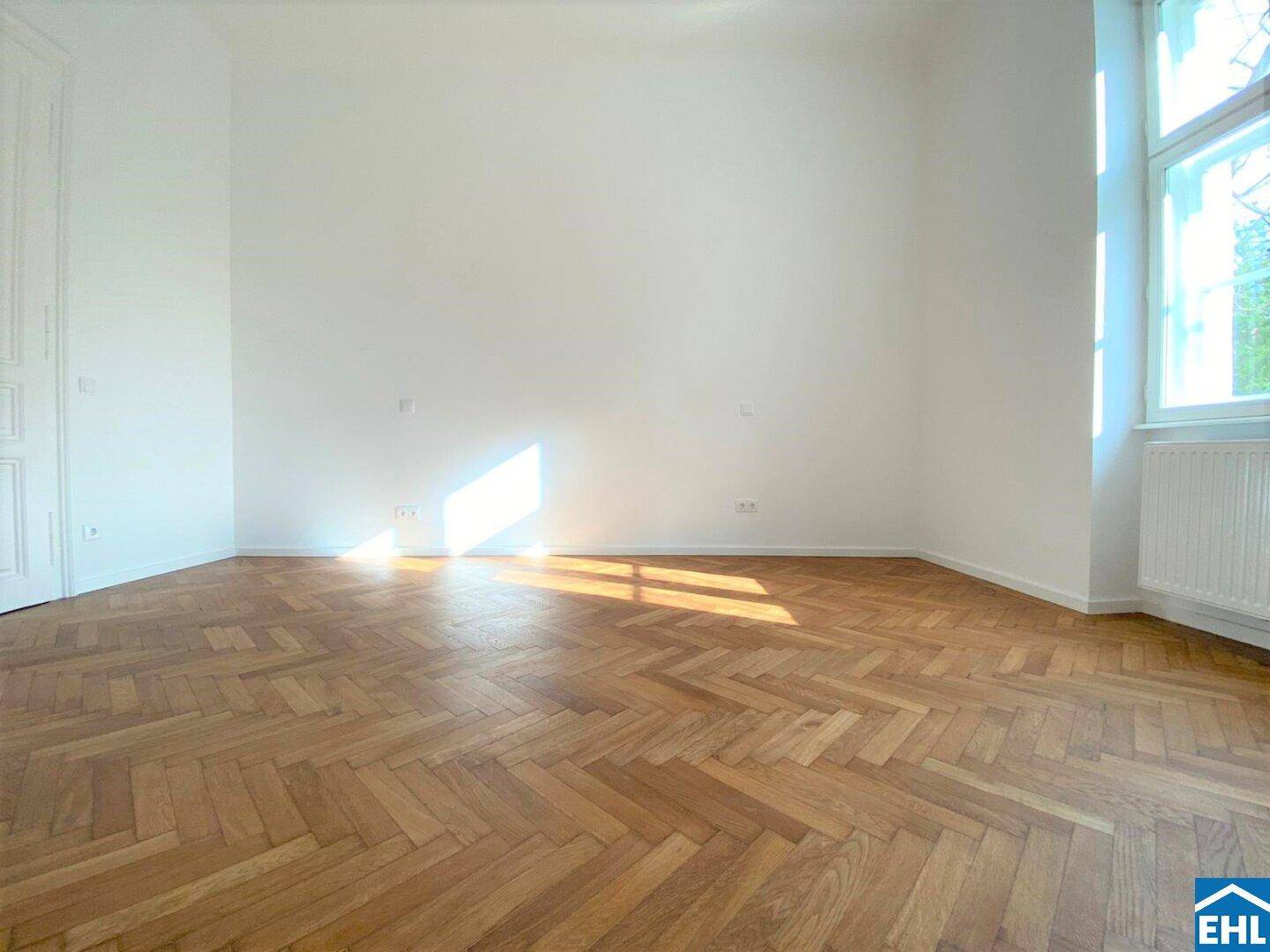 Großes, helles Zimmer mit klassischem Fischgrätparkett und hohen Decken, ideal für einen Wohnbereich.