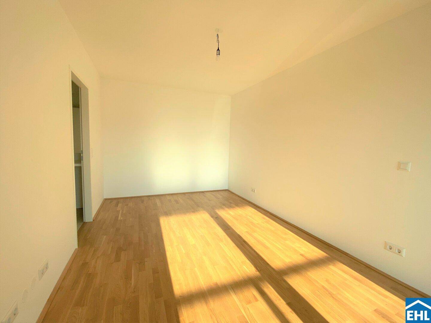 Leerer Raum mit hellem Holzfußboden und weißen Wänden, durchflutet von natürlichem Sonnenlicht.