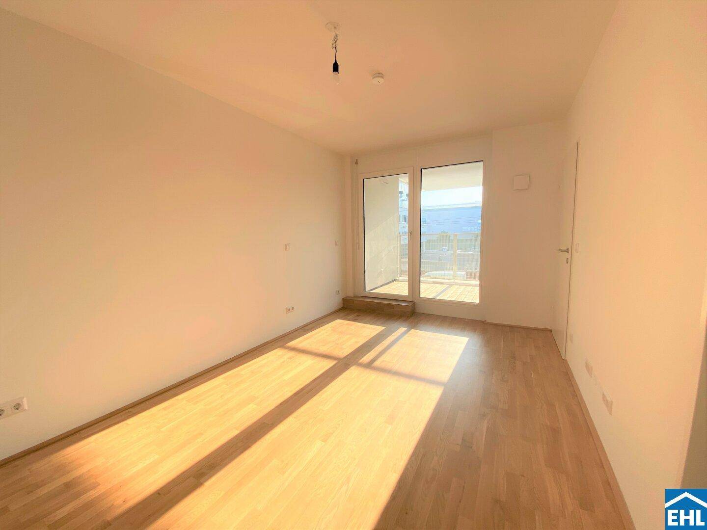 Heller, leerer Raum mit hellem Holzfußboden und großen Fenstern, die viel Sonnenlicht hereinlassen.