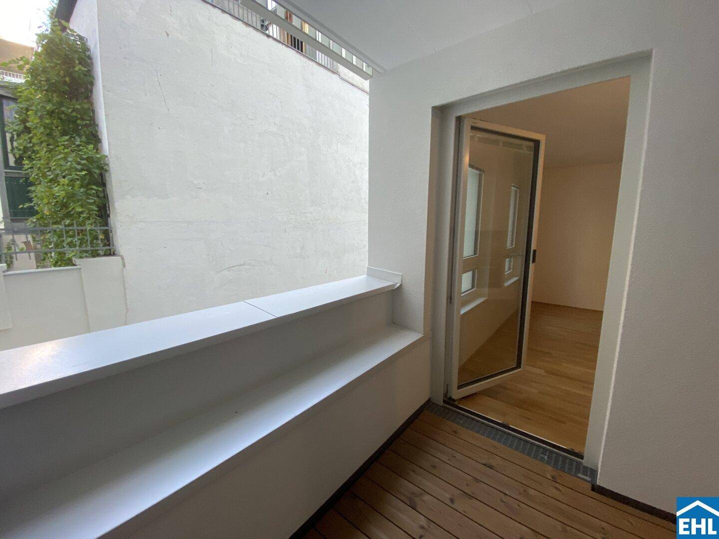 Gemütlicher Balkon mit Holzboden und Zugang zum Innenbereich, umgeben von grüner Bepflanzung.