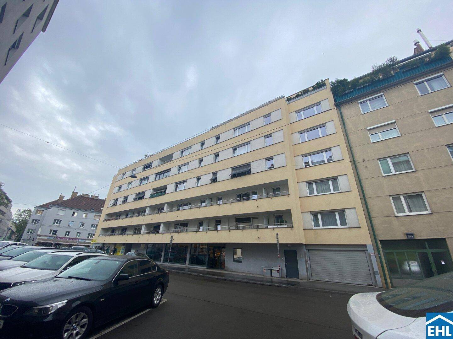 Mehrstöckiges Wohngebäude mit Geschäften im Erdgeschoss und parkenden Autos auf der Straße.