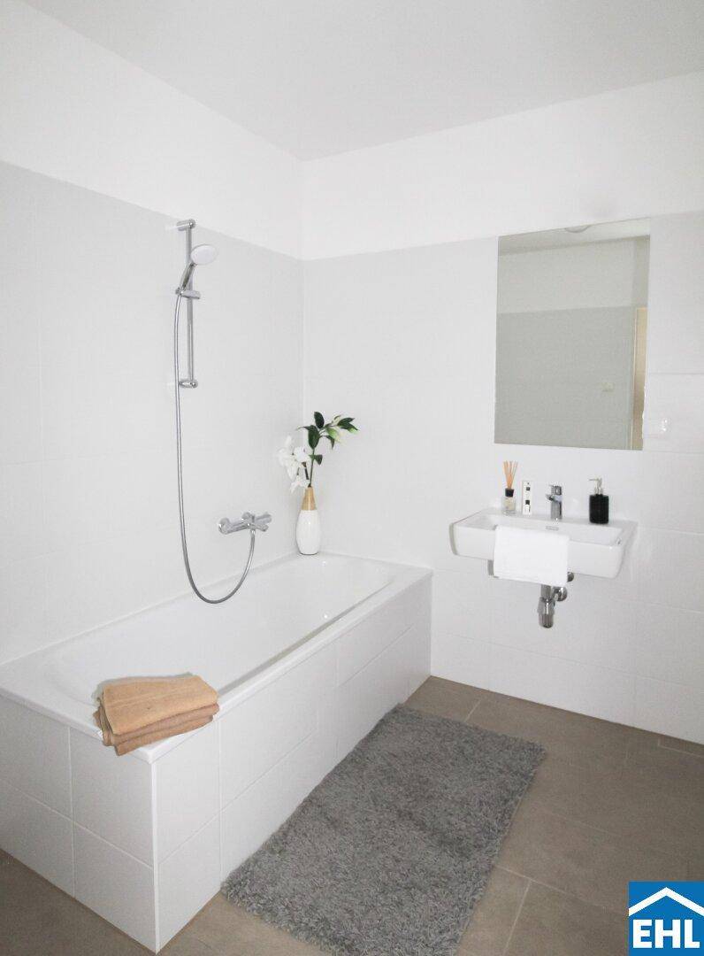 Helles Badezimmer mit Badewanne, Wandwaschbecken und großen weißen Fliesen.