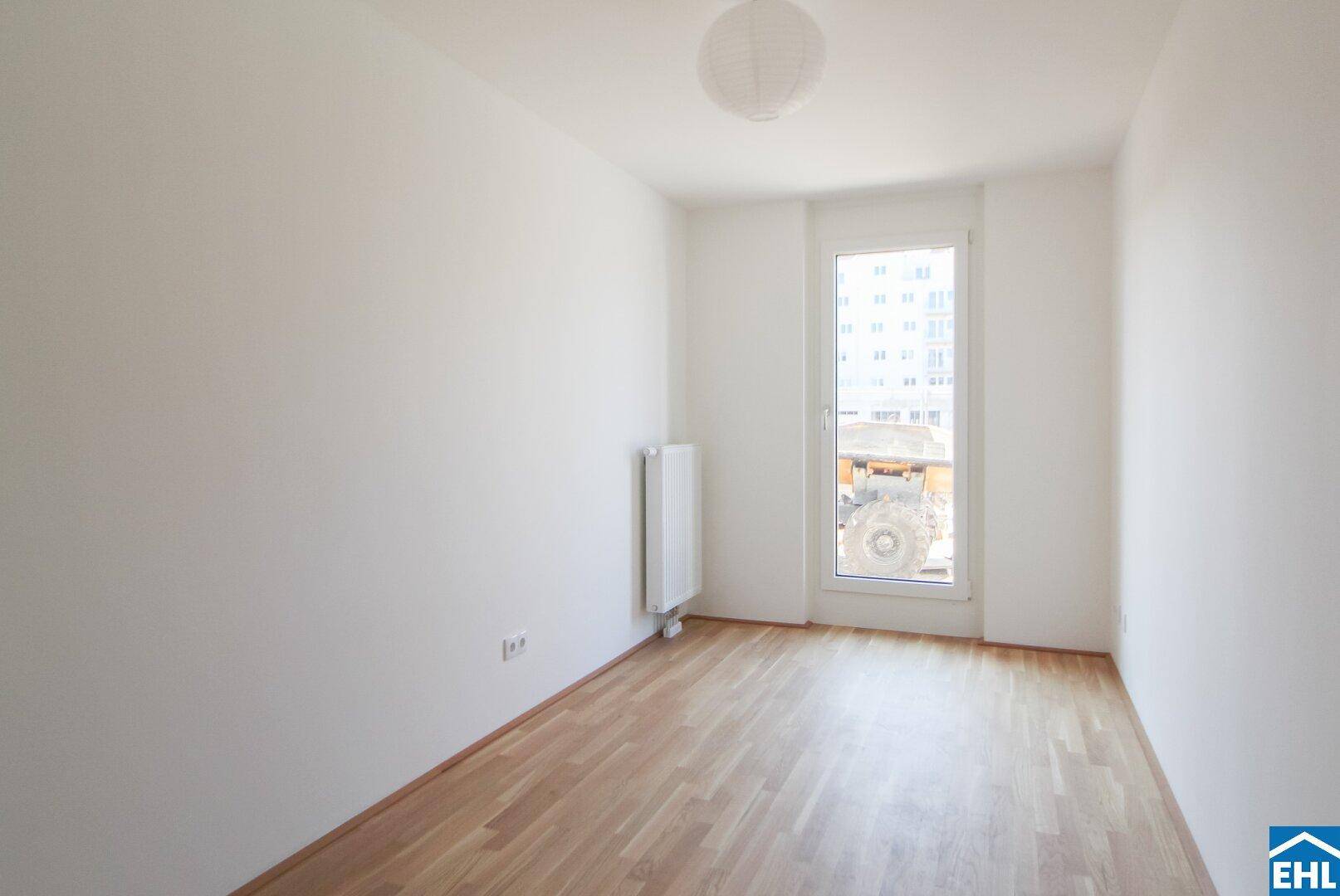 Leeres Zimmer mit Holzboden, großem Fenster und Heizkörper.