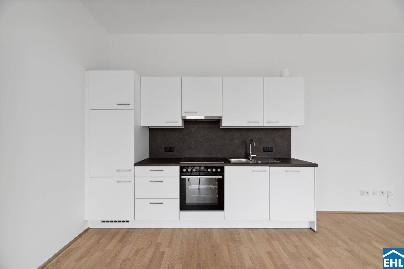 Moderne Einbauküche mit weißen Fronten, dunkler Arbeitsplatte und integriertem Backofen in einem hellen Raum.
