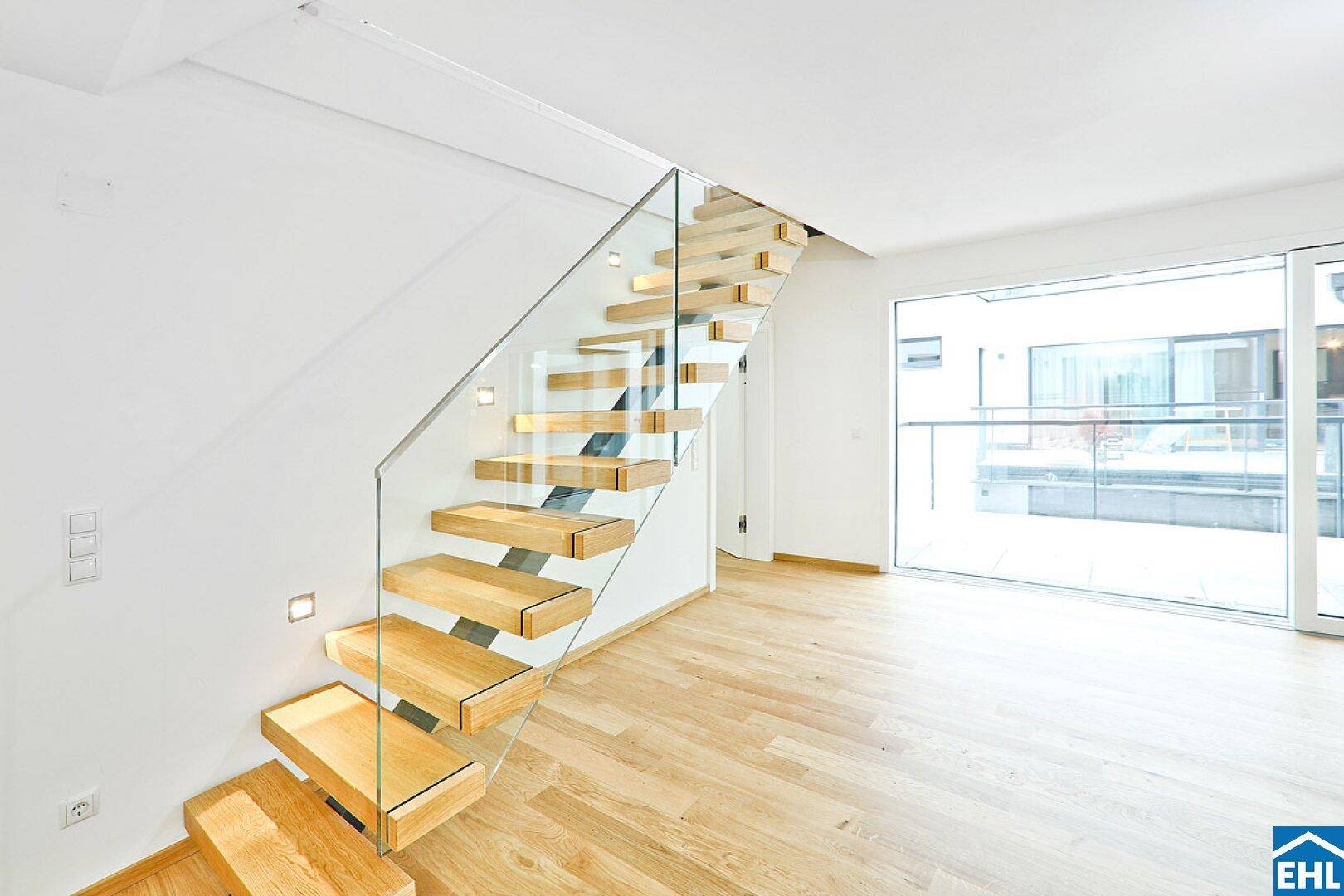 Elegante Holztreppe mit Glasgeländer in einem hellen Wohnbereich mit großen Fenstern.
