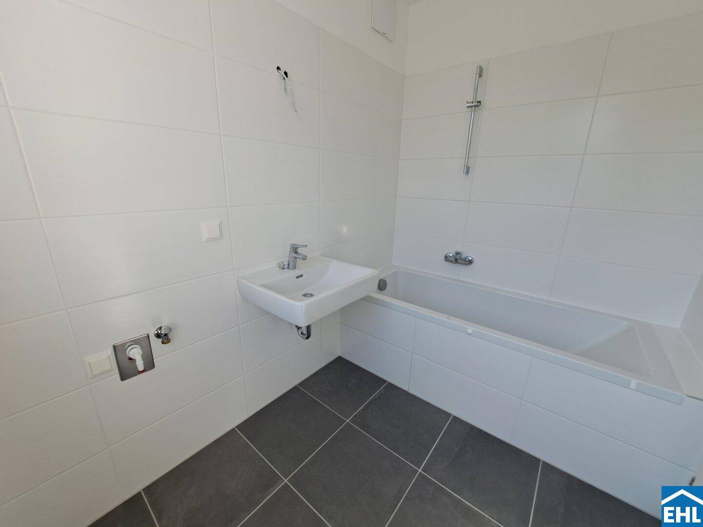 Modernes Badezimmer mit weißen Fliesen, dunklem Boden, Waschbecken und Badewanne mit Duschfunktion.