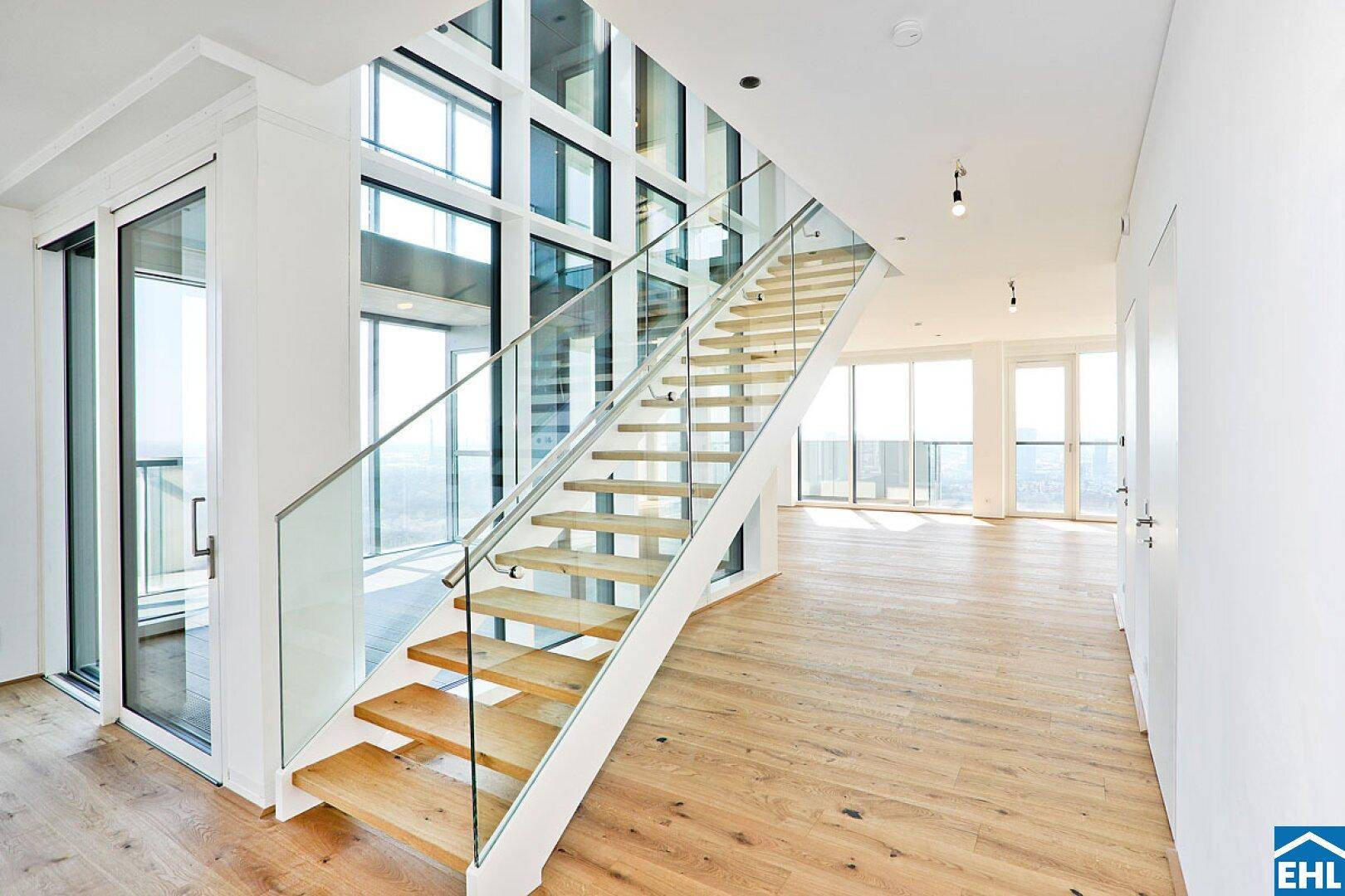 Moderne Holztreppe mit Glasgeländer in einem hellen, offenen Wohnbereich mit weitem Ausblick.