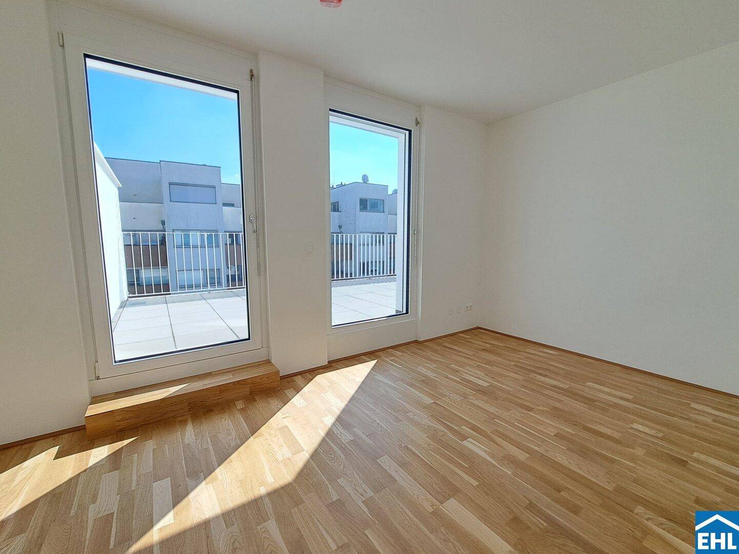 Helles, leeres Zimmer mit Holzboden und großen Fenstern, die auf eine Terrasse blicken.