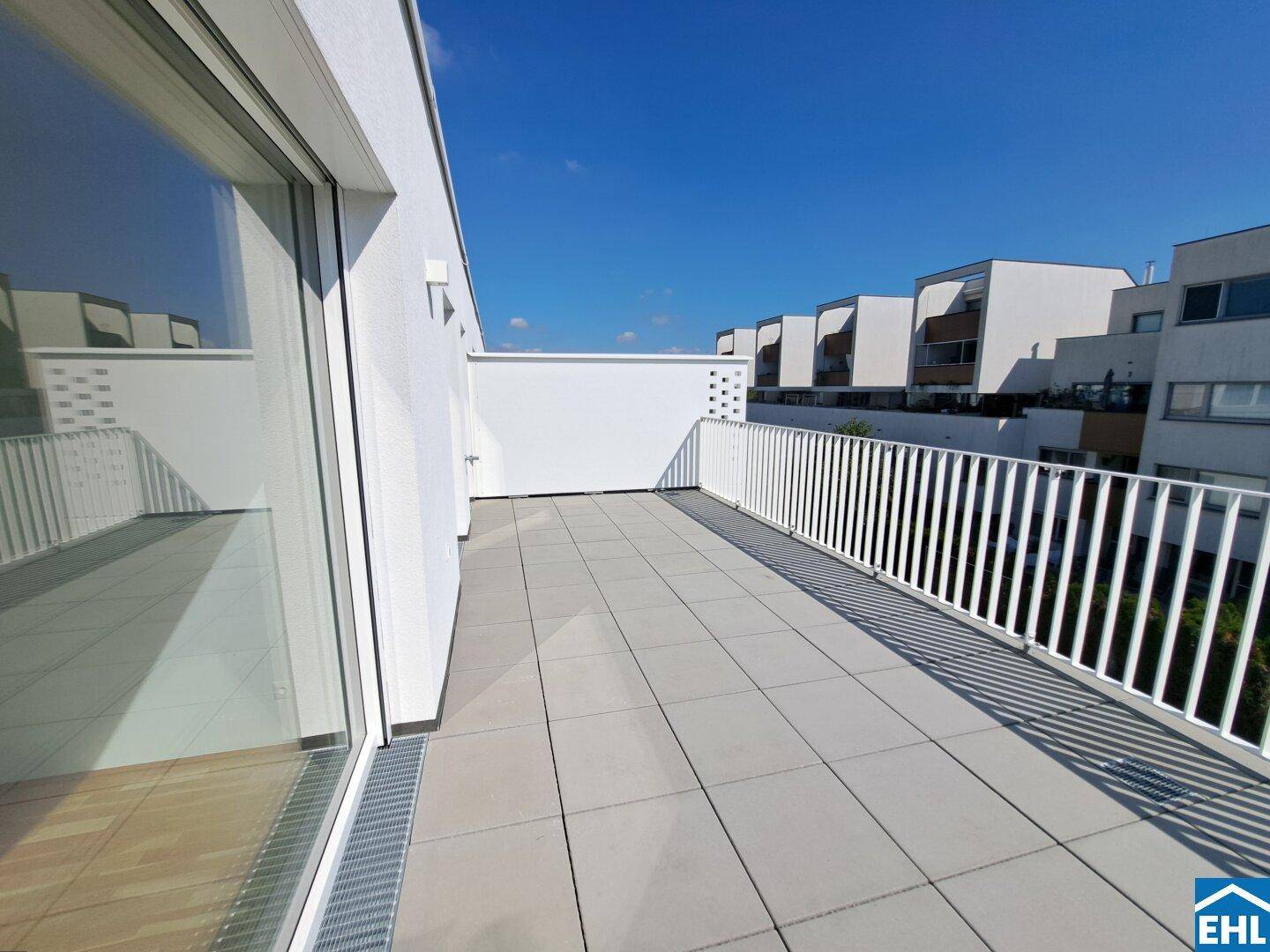 Geräumige Dachterrasse mit hellen Fliesen und weißem Geländer, umgeben von Neubauten.
